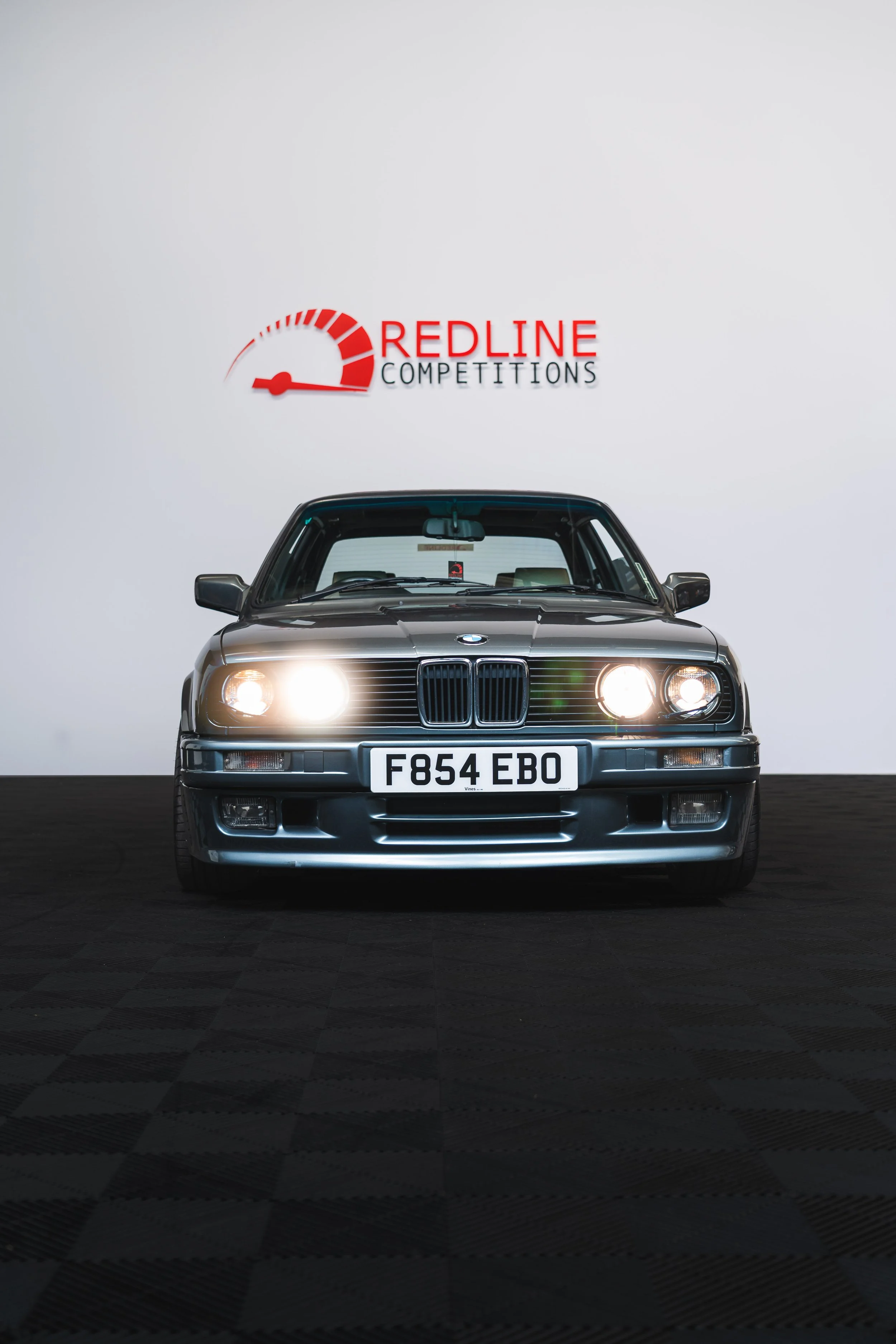 A black BMW car with headlights on, facing forward, in front of a white wall with a red and black 'Redline Competitions' logo.