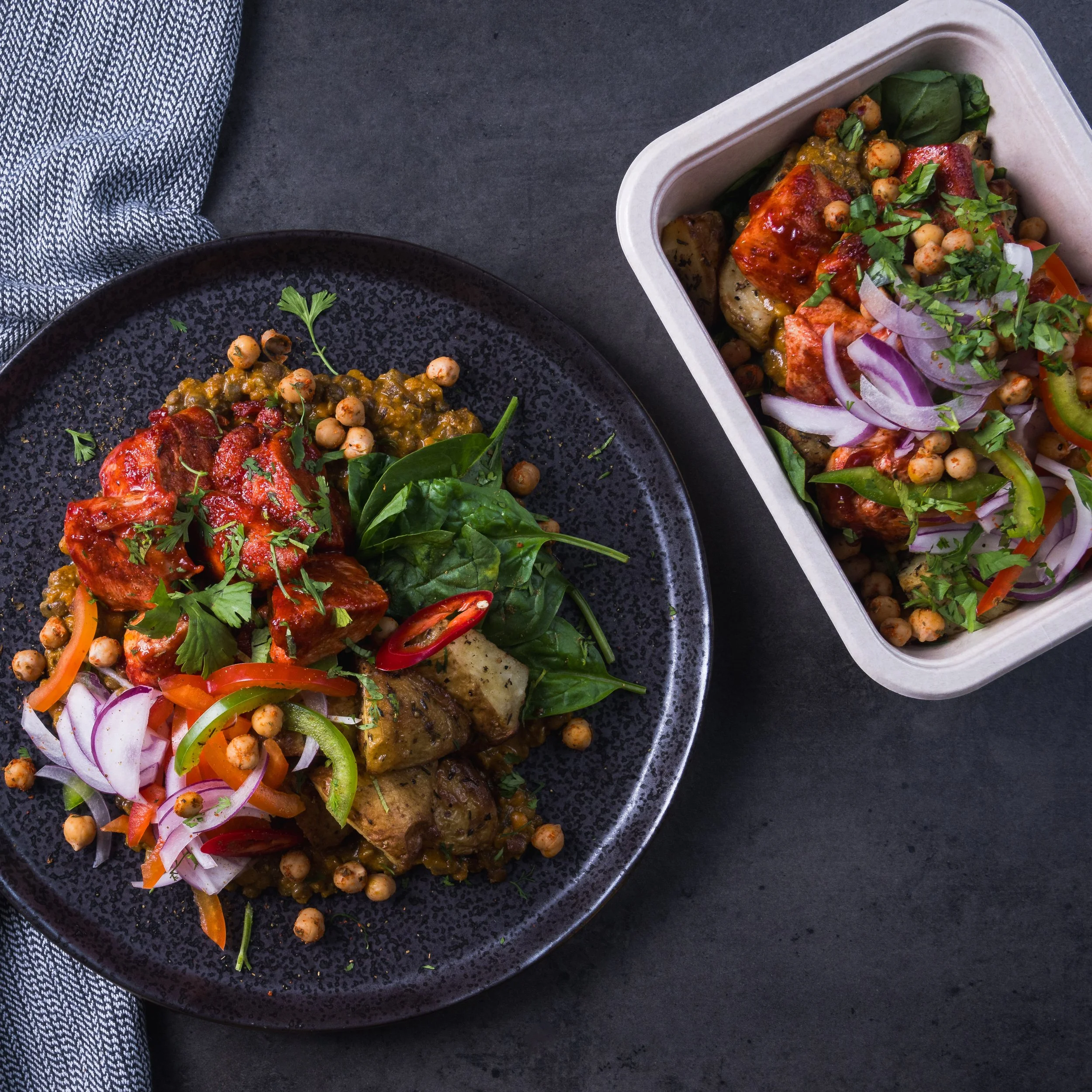 Plated Indian curry with vegetables and chickpeas, served with fresh spinach and garnished with cilantro and sliced red chilies, along with a similar dish in a rectangular container.