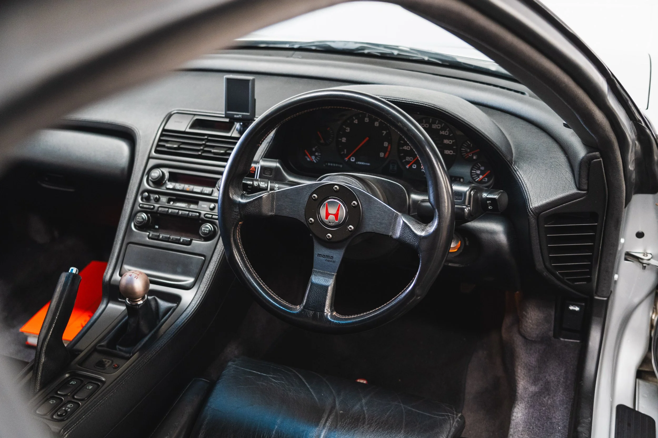 Interior of a sports car focusing on the steering wheel with a Honda emblem, manual gear shift, dashboard gauges, and center console with radio and controls.