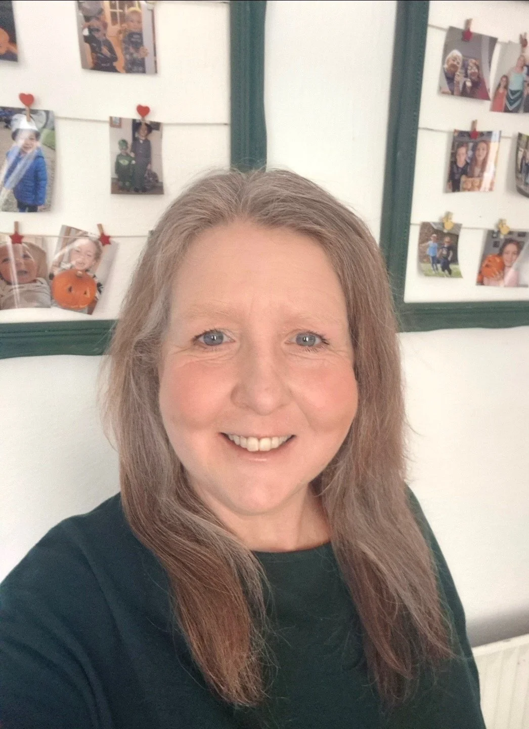 A woman with long gray-blonde hair smiling for a selfie, with a photo display in the background showing various pictures of children and people, some decorated with small red hearts.