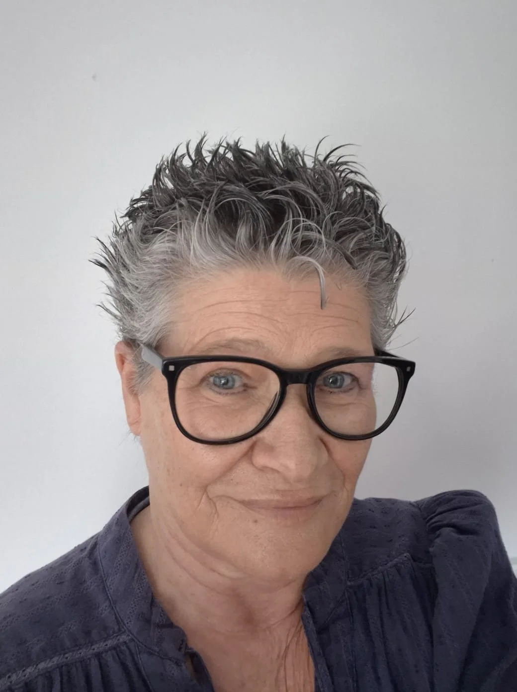 Close-up of a smiling woman with short, spiky, salt-and-pepper hair, wearing large black glasses, against a plain white background.