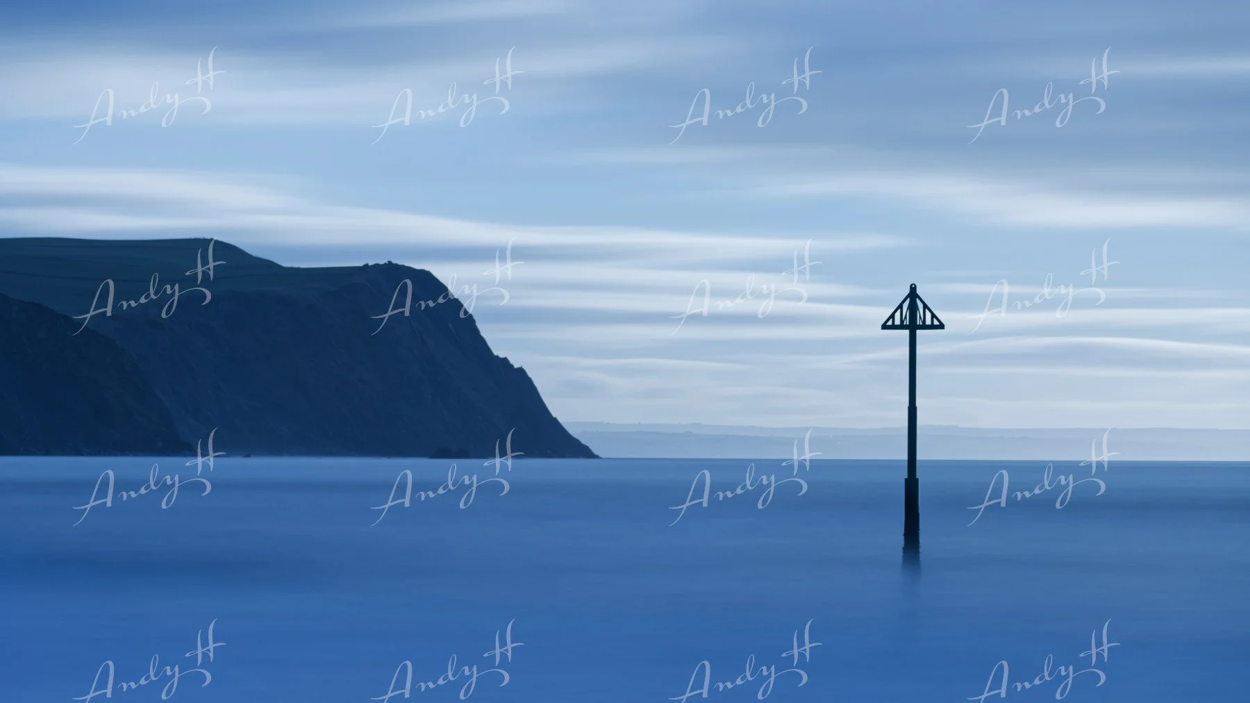 A seascape fine art print, A long exposure photograph of a seascape showing a calm, blue ocean with a distant cliff on the left and a solitary navigation marker or buoy in the water toward the right, under a cloudy sky.