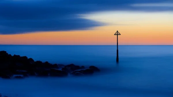 A seascape fine art print, Calm ocean at sunset with dark rocks in foreground and a navigational beacon in the water. The sky features a gradient of colors from dark blue to orange near the horizon.