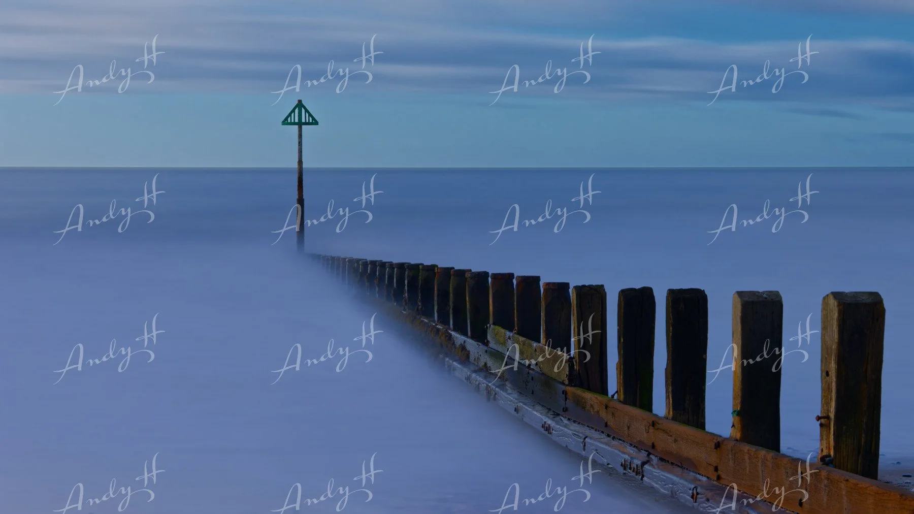 A seascape fine art print, A wooden breakwater extending into the calm ocean, with a navigational marker at the end, under a cloudy sky.