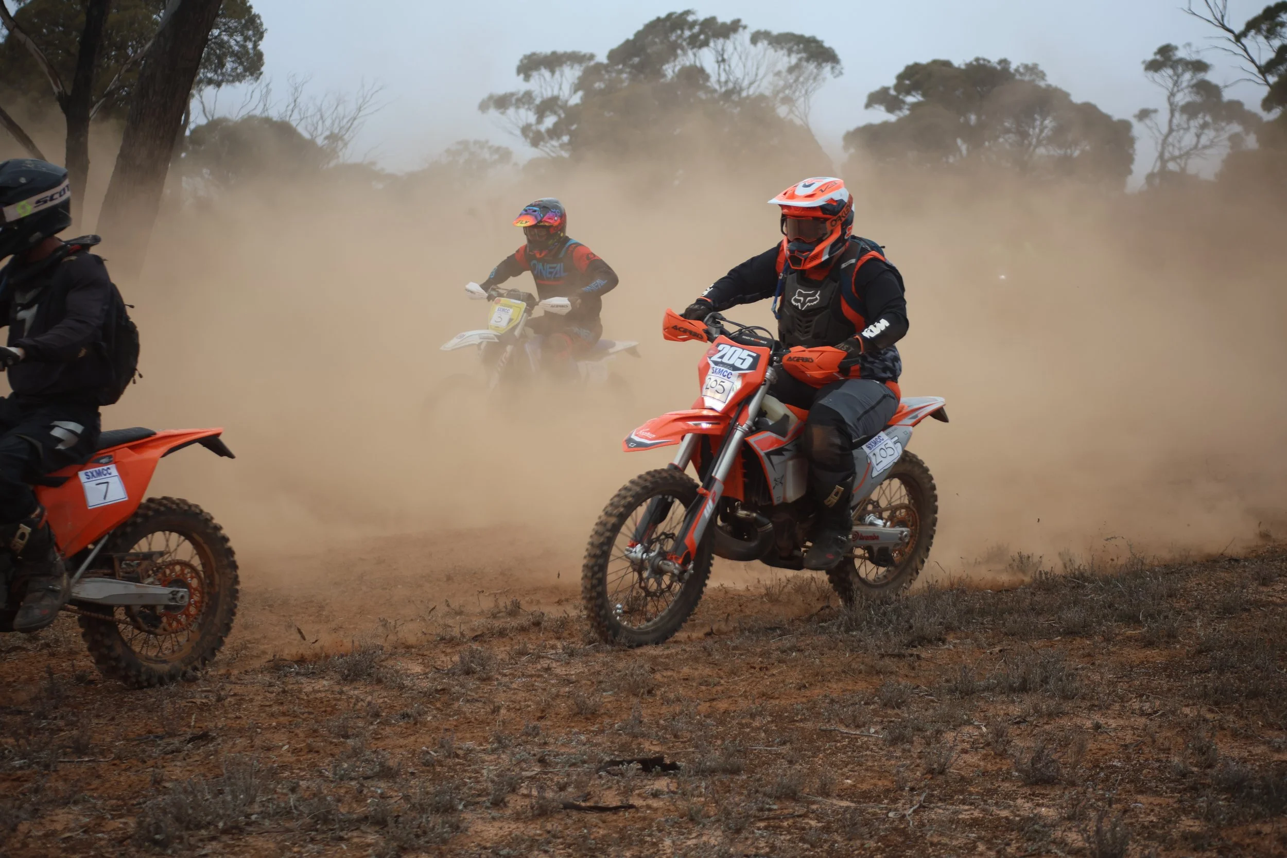 Group of off-road motorcyclists riding on a dirt trail, kicking up dust, with trees in the background.