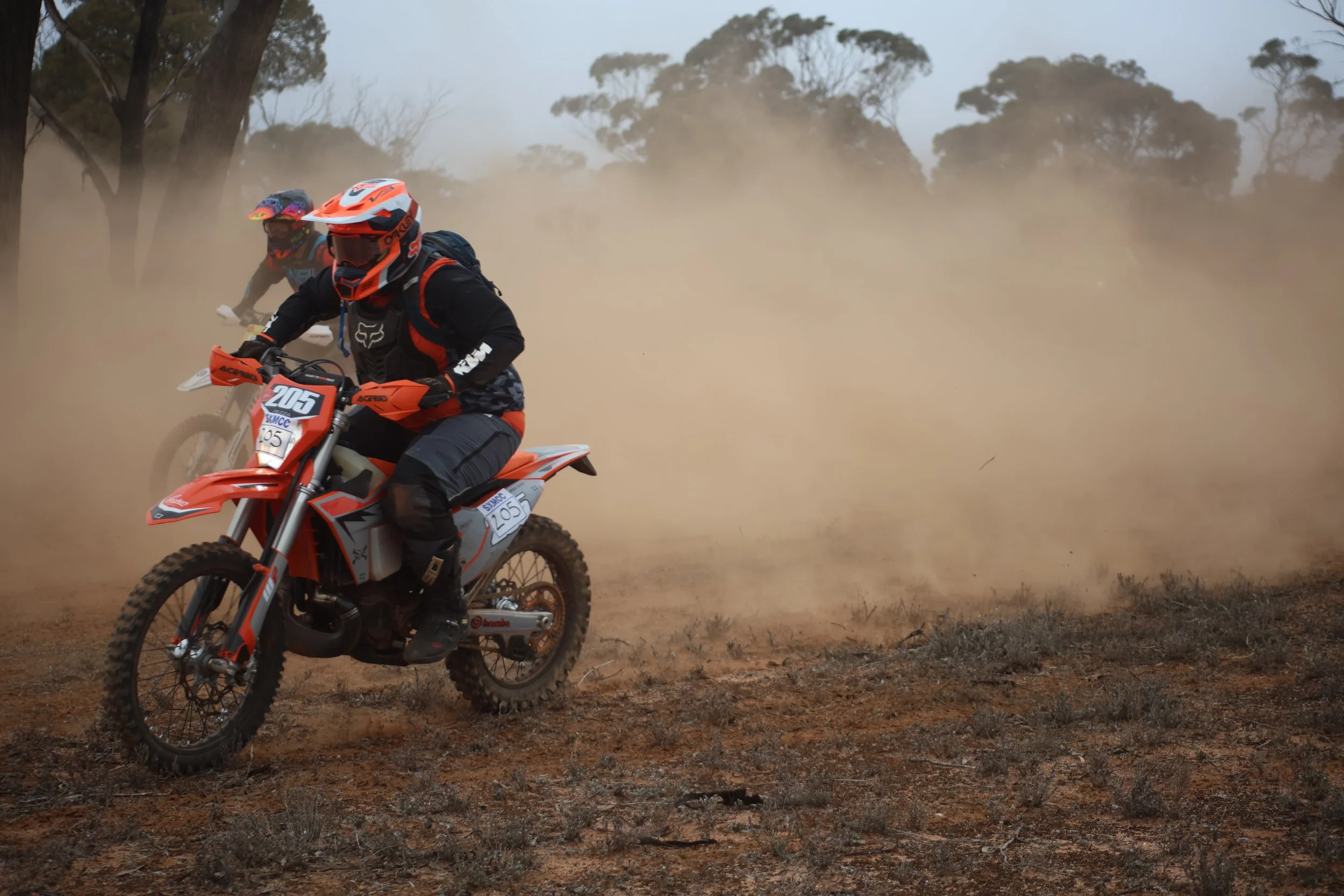 Two motocross riders riding on a dirt trail through a dusty, open landscape with sparse vegetation and trees in the background, wearing helmets and protective gear.