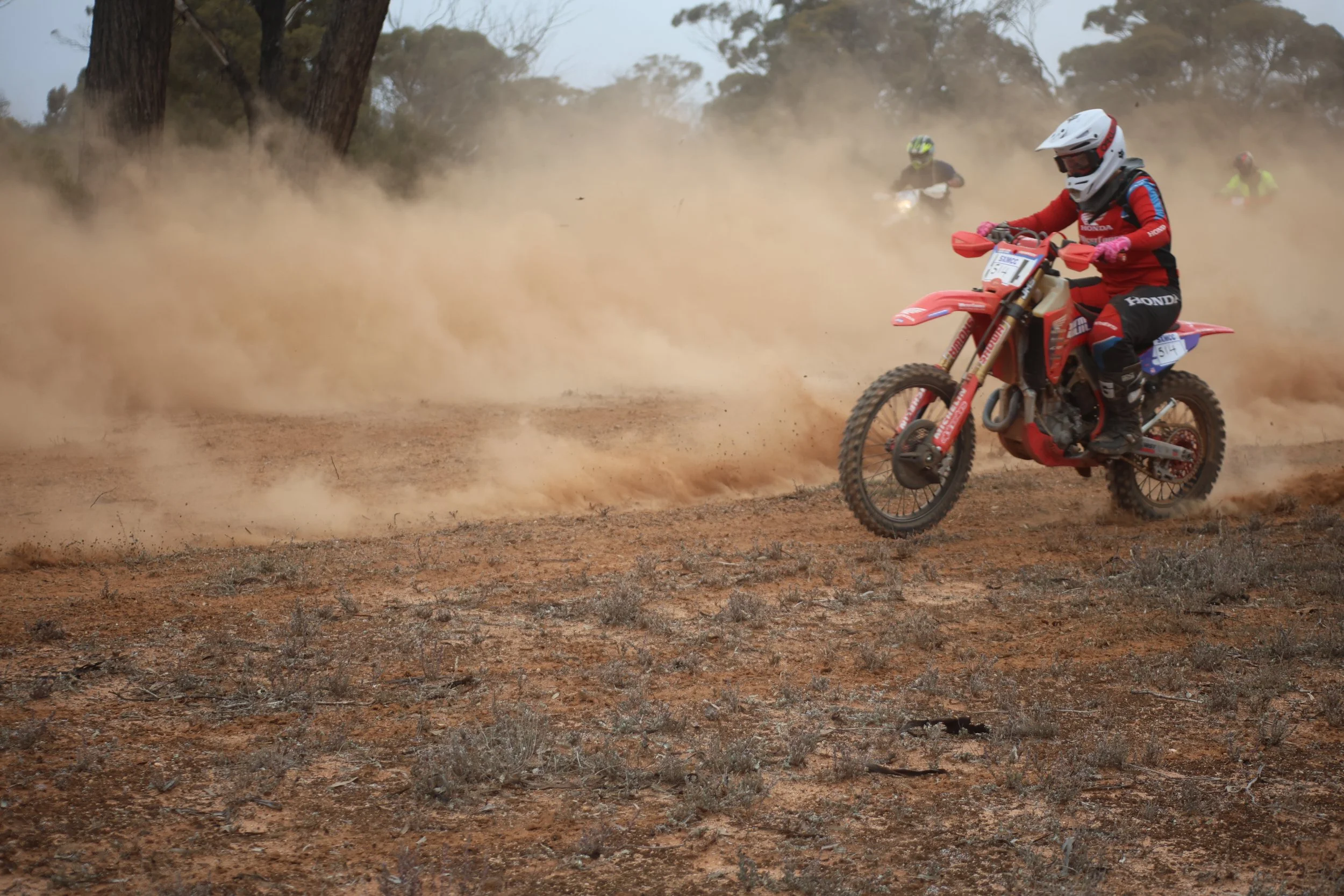 A person riding a red dirt bike through a dusty, dry landscape with trees in the background, wearing a white helmet and red and blue racing gear.