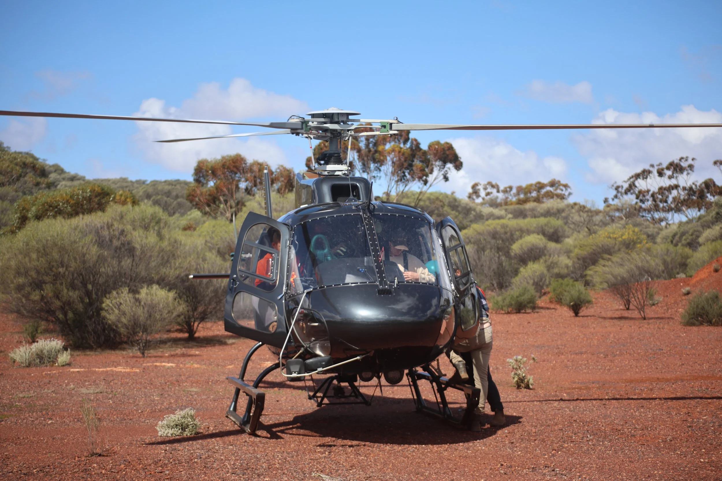 A black helicopter landing on a reddish dirt field with desert shrubs and trees in the background under a partly cloudy sky.