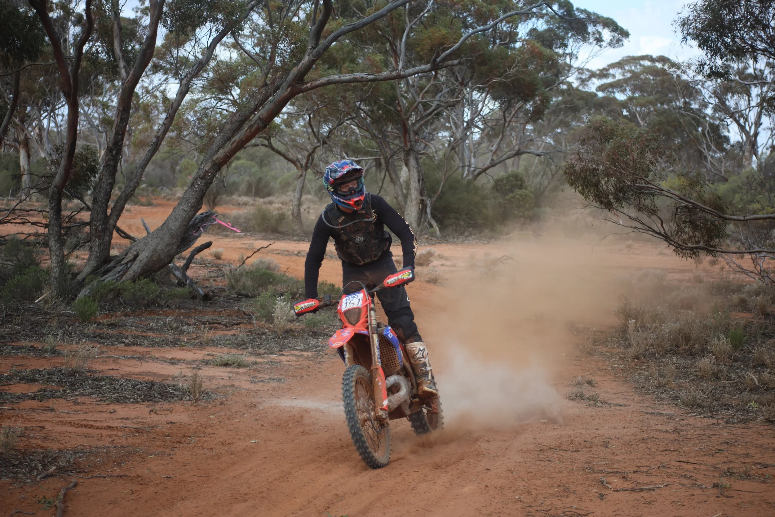 A person riding a dirt bike on a dusty trail through a wooded area with trees and dry bushes.