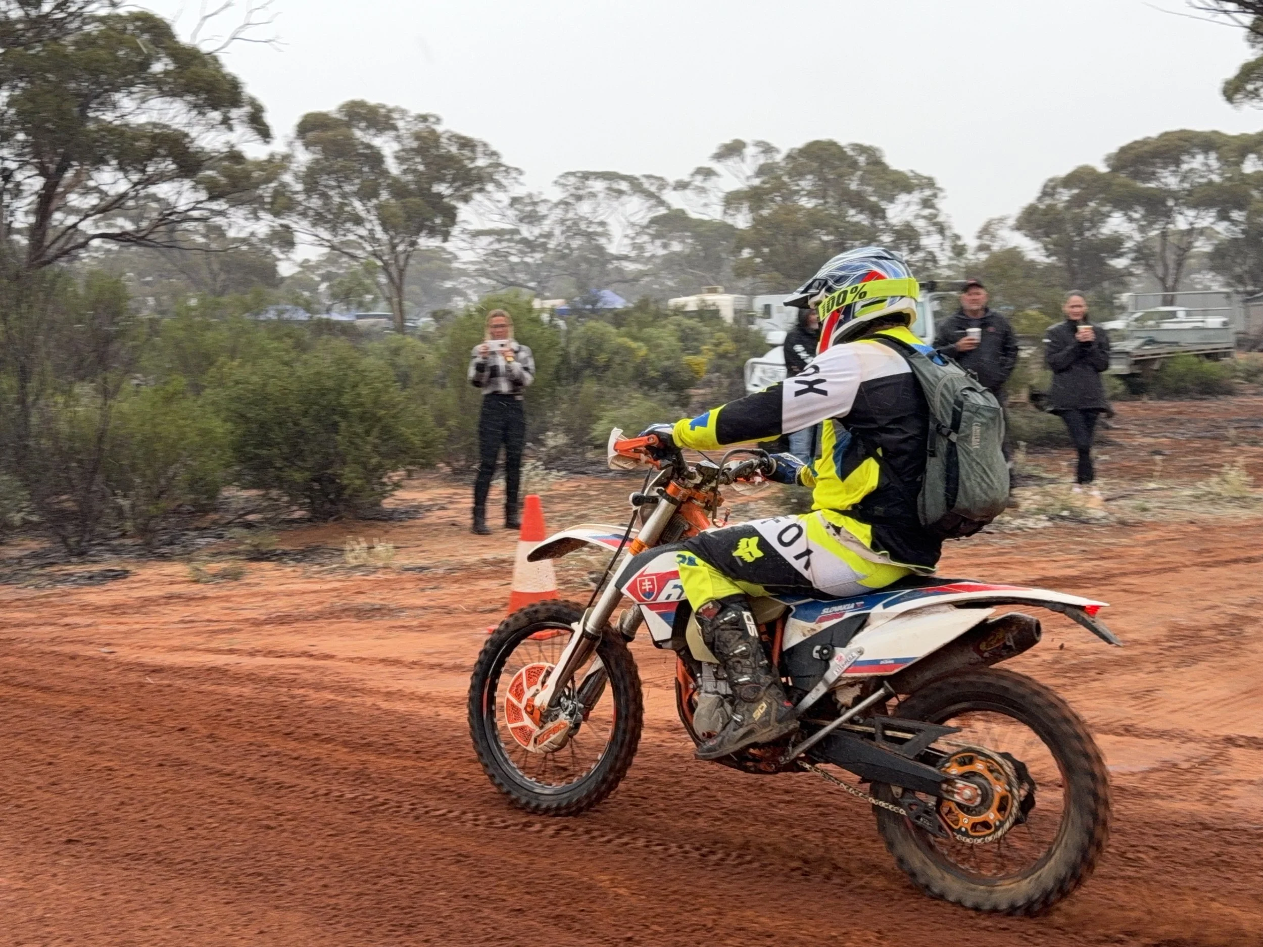 A person in motocross gear riding a dirt bike on a trail, with four spectators taking photos or videos in a wooded area.