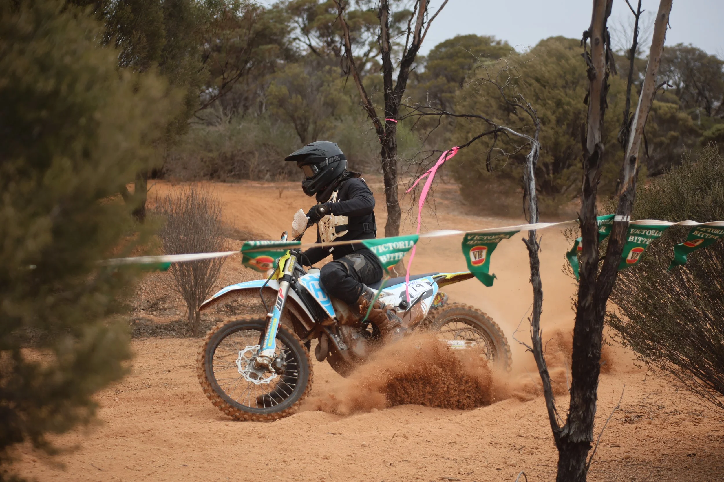 A motocross rider wearing a black helmet and riding gear navigates a dirt trail through a desert landscape with sparse bushes and trees. The rider kicks up a cloud of dust as they turn, and temporary barriers and green flags mark the race course.
