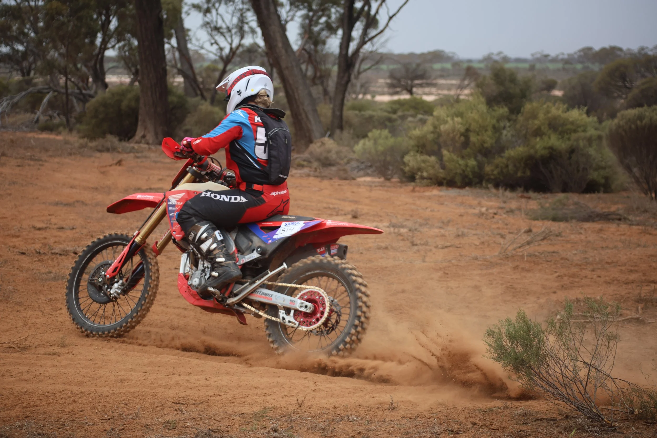 A motocross rider on a red dirt bike, wearing a helmet and protective gear, riding on a dirt trail through a semi-arid landscape with sparse vegetation and trees.