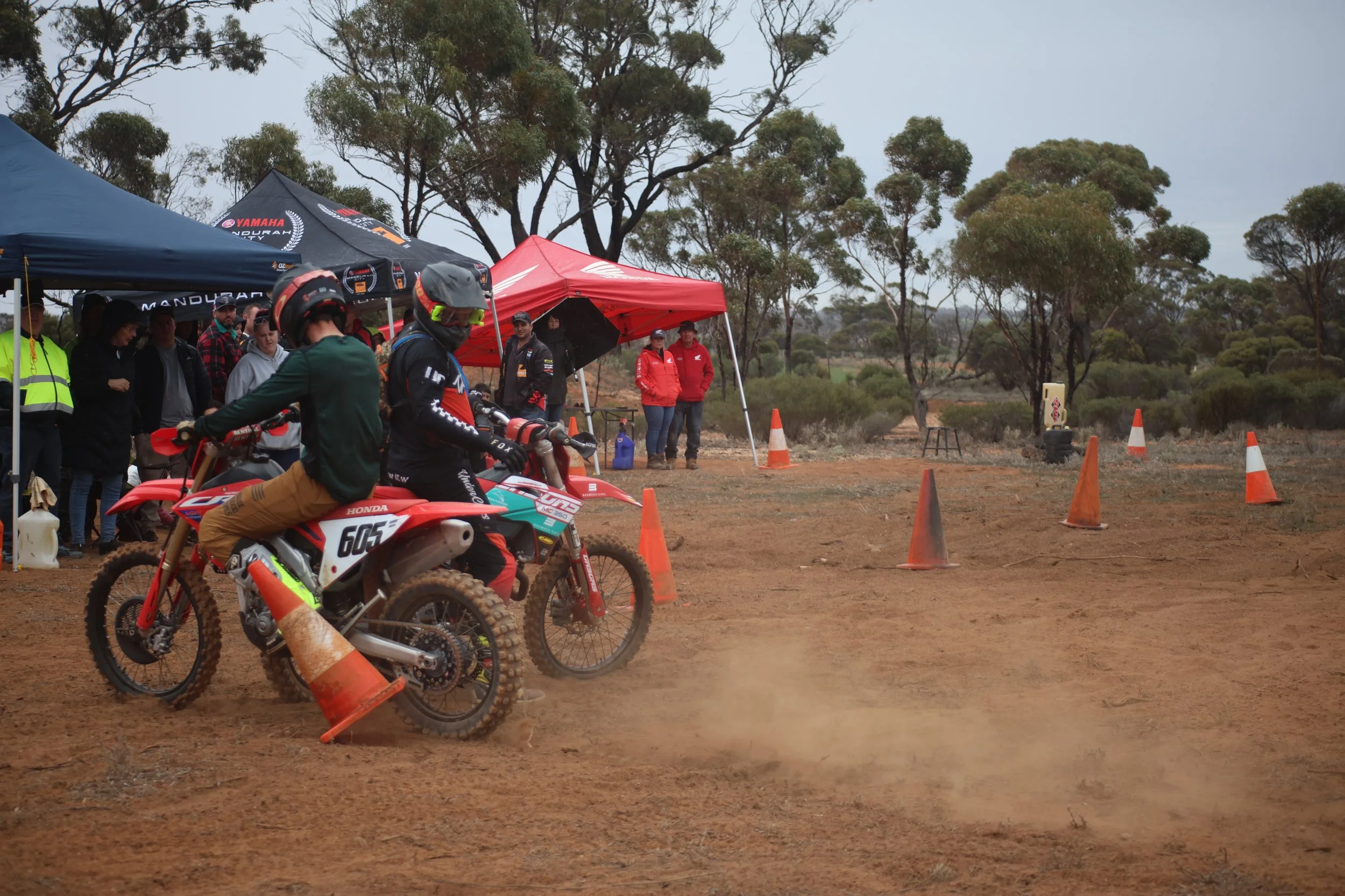 Two motocross riders on dirt bikes at the starting line, with a crowd of spectators and tents in the background, dirt track with orange cones, in a rural outdoor setting.