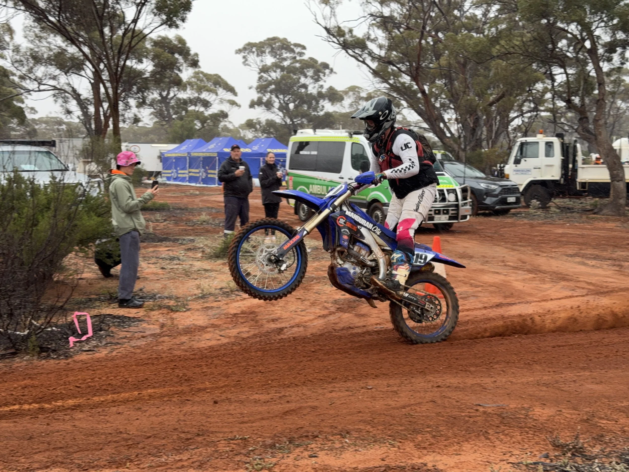 Motocross rider jumping on a dirt track, with spectators and emergency vehicles in the background.