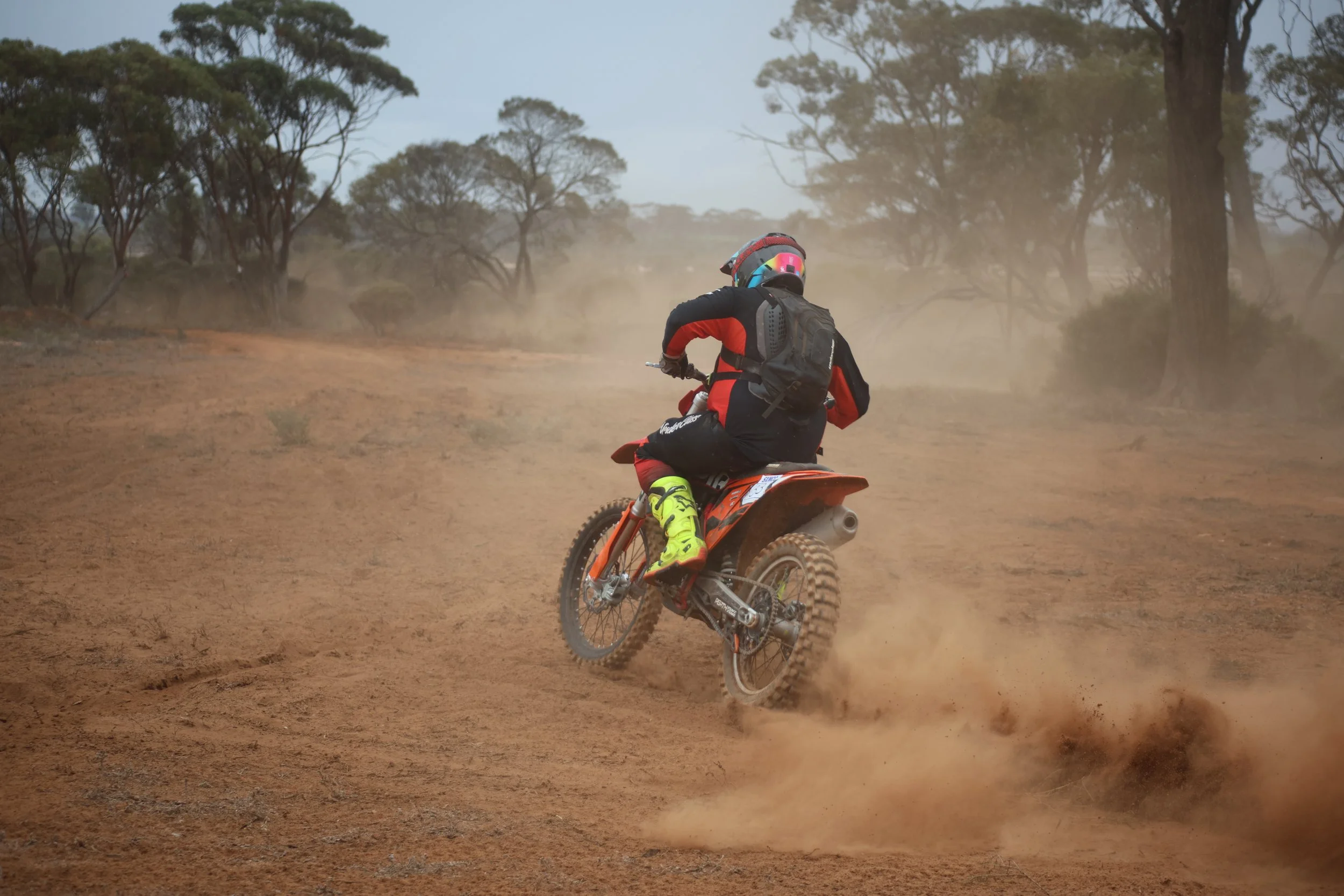 Dirt bike rider wearing a black and red suit with yellow boots riding on a dusty trail in a dry, wooded area.