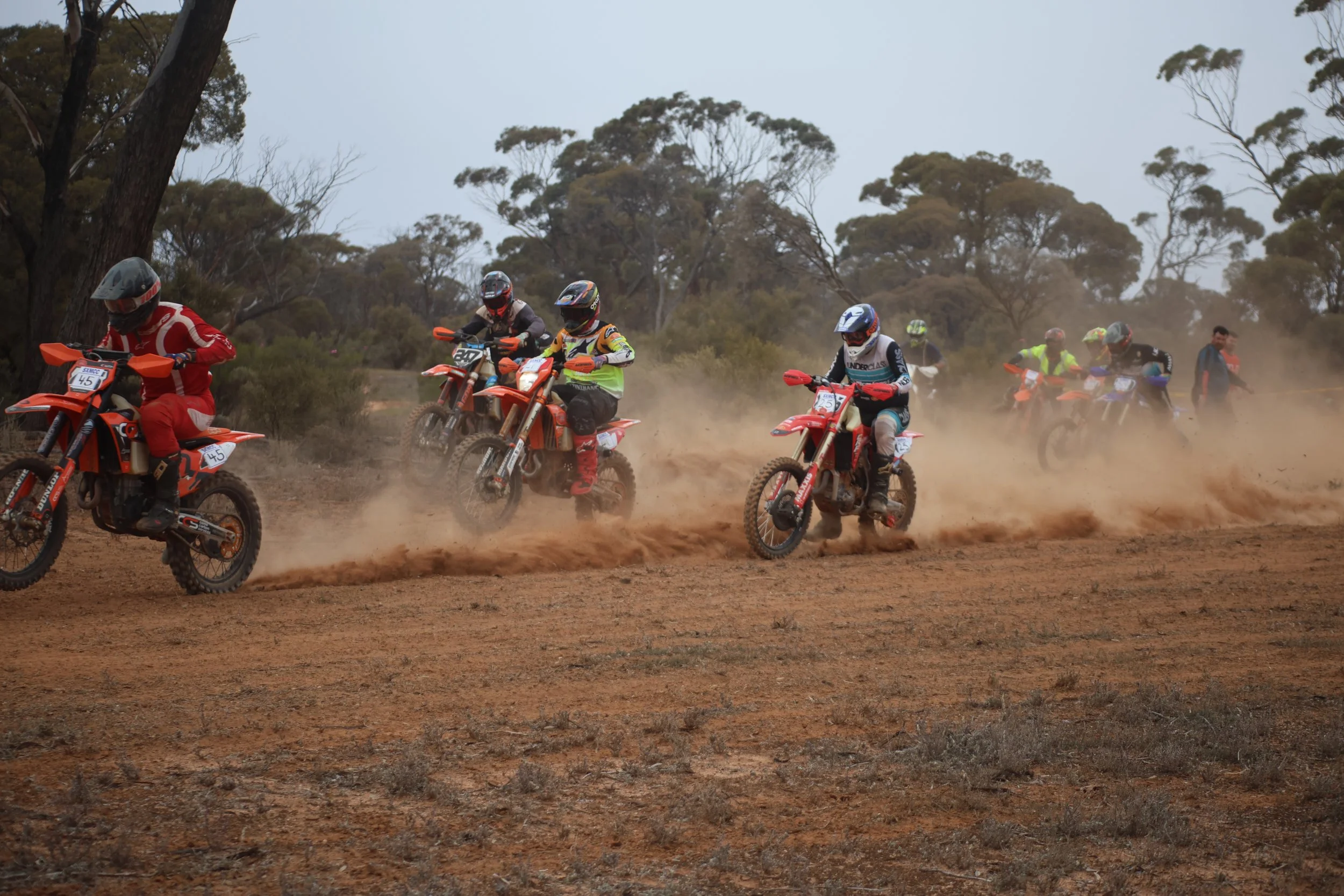 Group of motocross riders racing on a dirt track with dust clouds, wearing helmets and protective gear, with trees in the background.
