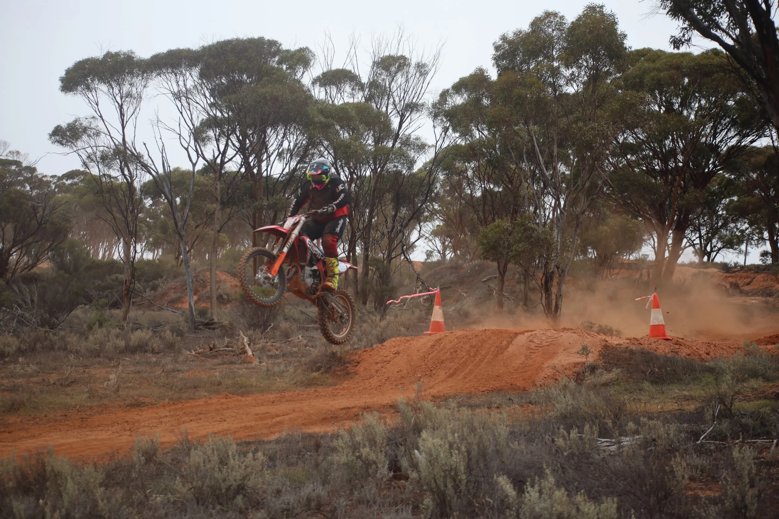 Motocross rider in black and red gear airborne over a dirt jump in a dry, wooded landscape with trees and dust clouds.