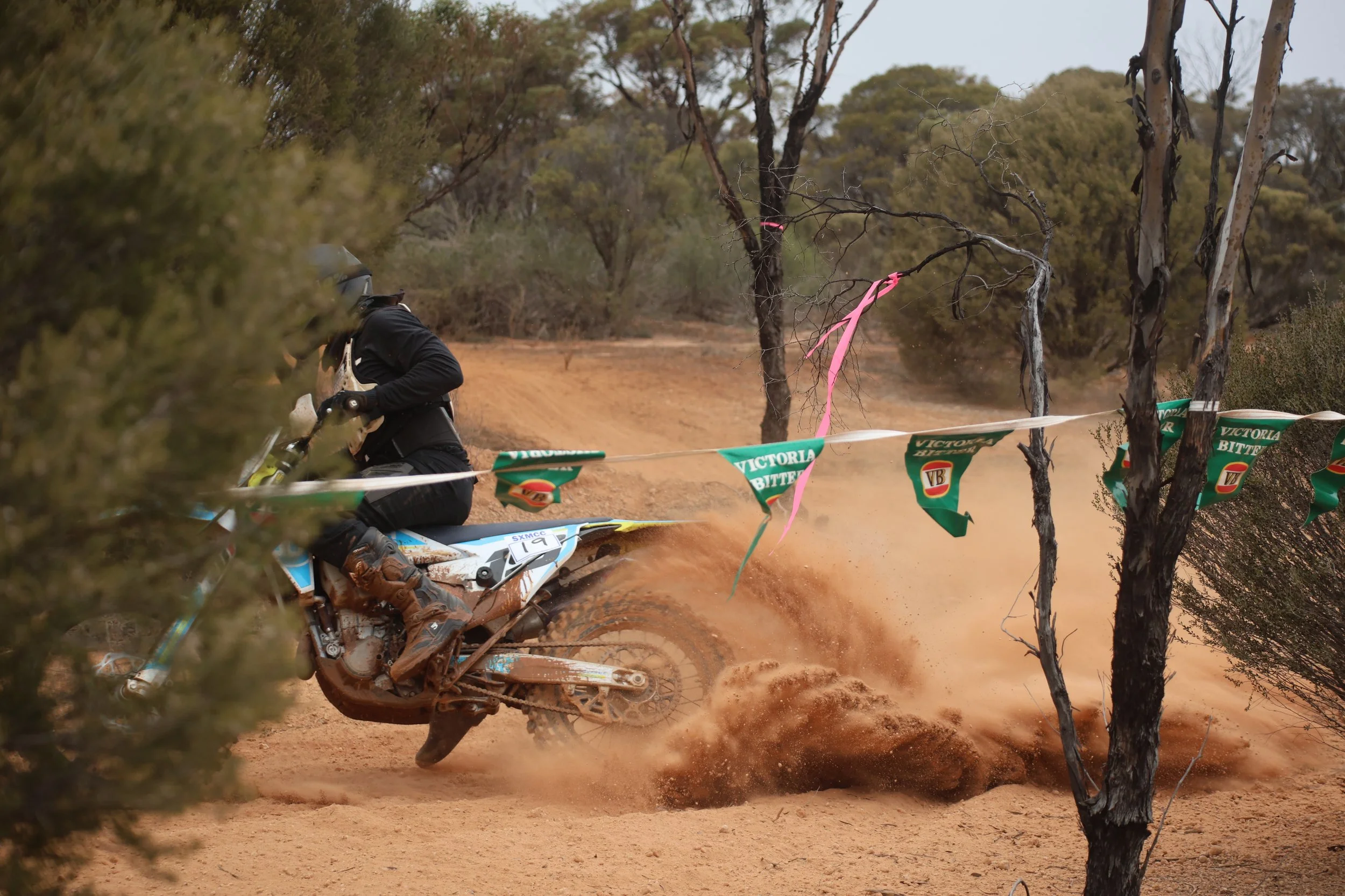 Motocross rider wearing helmet and riding gear on a dirt bike kicking up dust on a trail with trees and bushes nearby, part of a race marked by flags and banners.