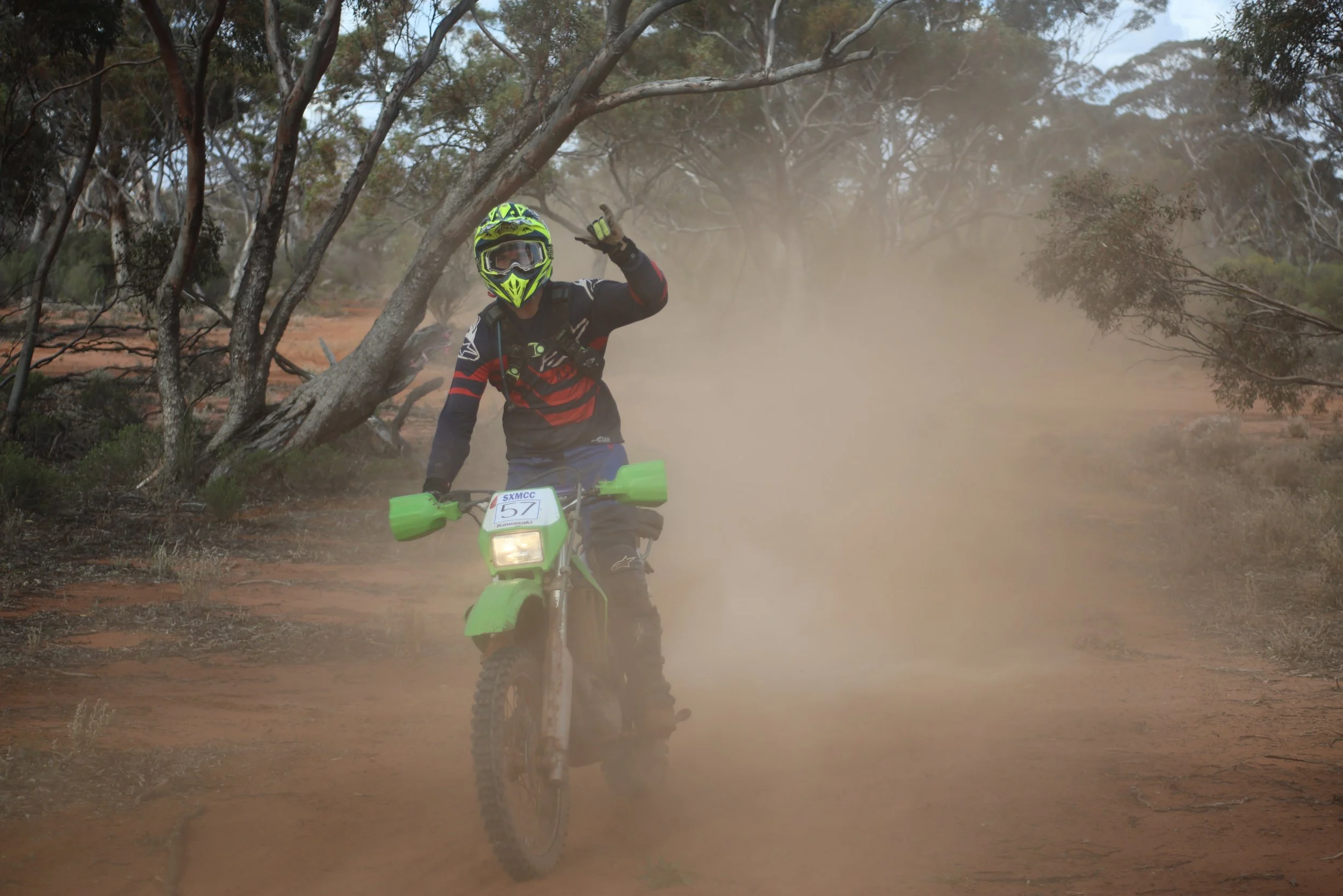 A person riding a green dirt bike on a dusty off-road trail surrounded by trees