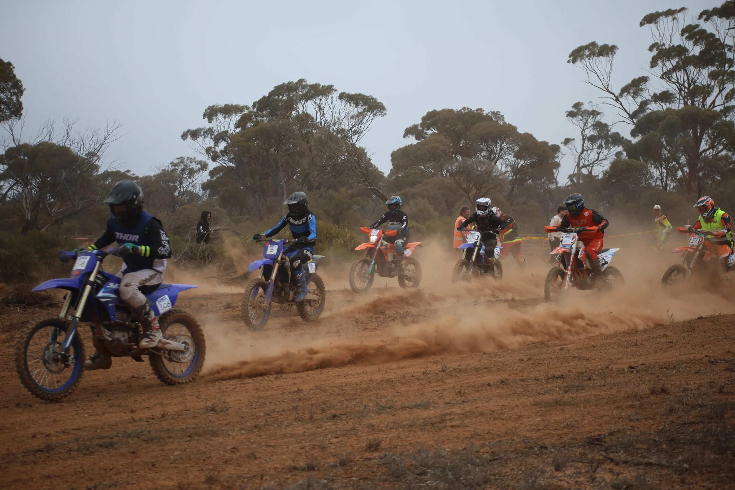 Motocross riders racing on a dirt track kicking up dust with trees in the background.