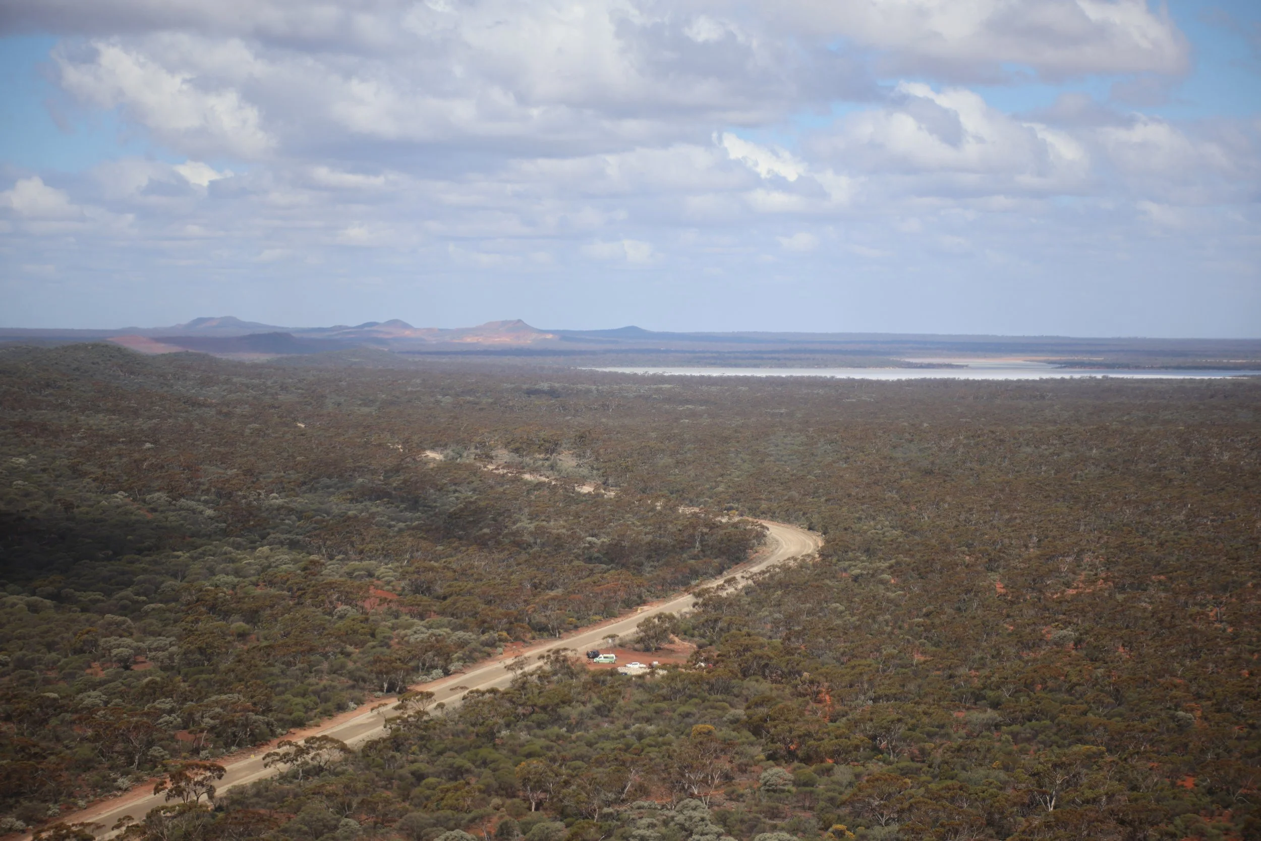 Aerial view of a dirt road winding through a vast, semi-arid landscape with scattered trees and shrubs, with hills and water bodies in the distance under a partly cloudy sky.