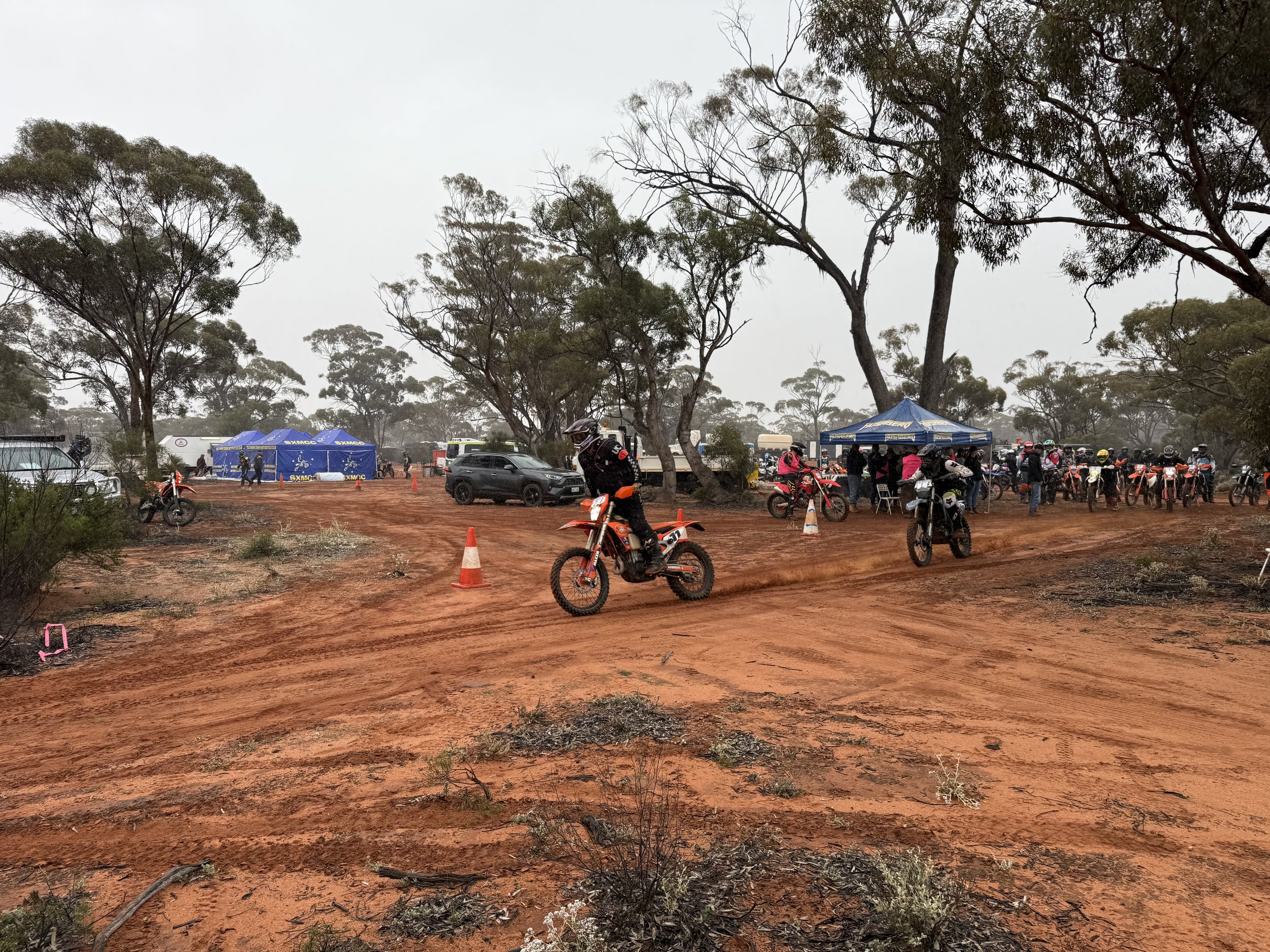 Off-road area with dirt ground, trees, motorcycle riders, and tents, possibly at a motocross or dirt bike event.