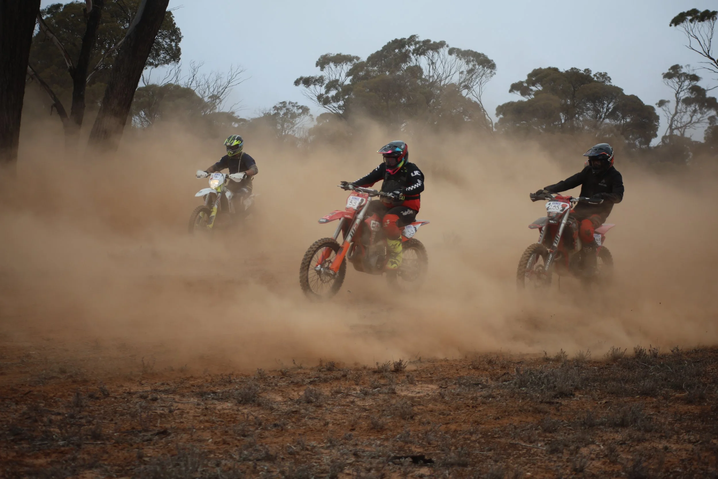 Three motocross riders riding through a dusty terrain with trees in the background.