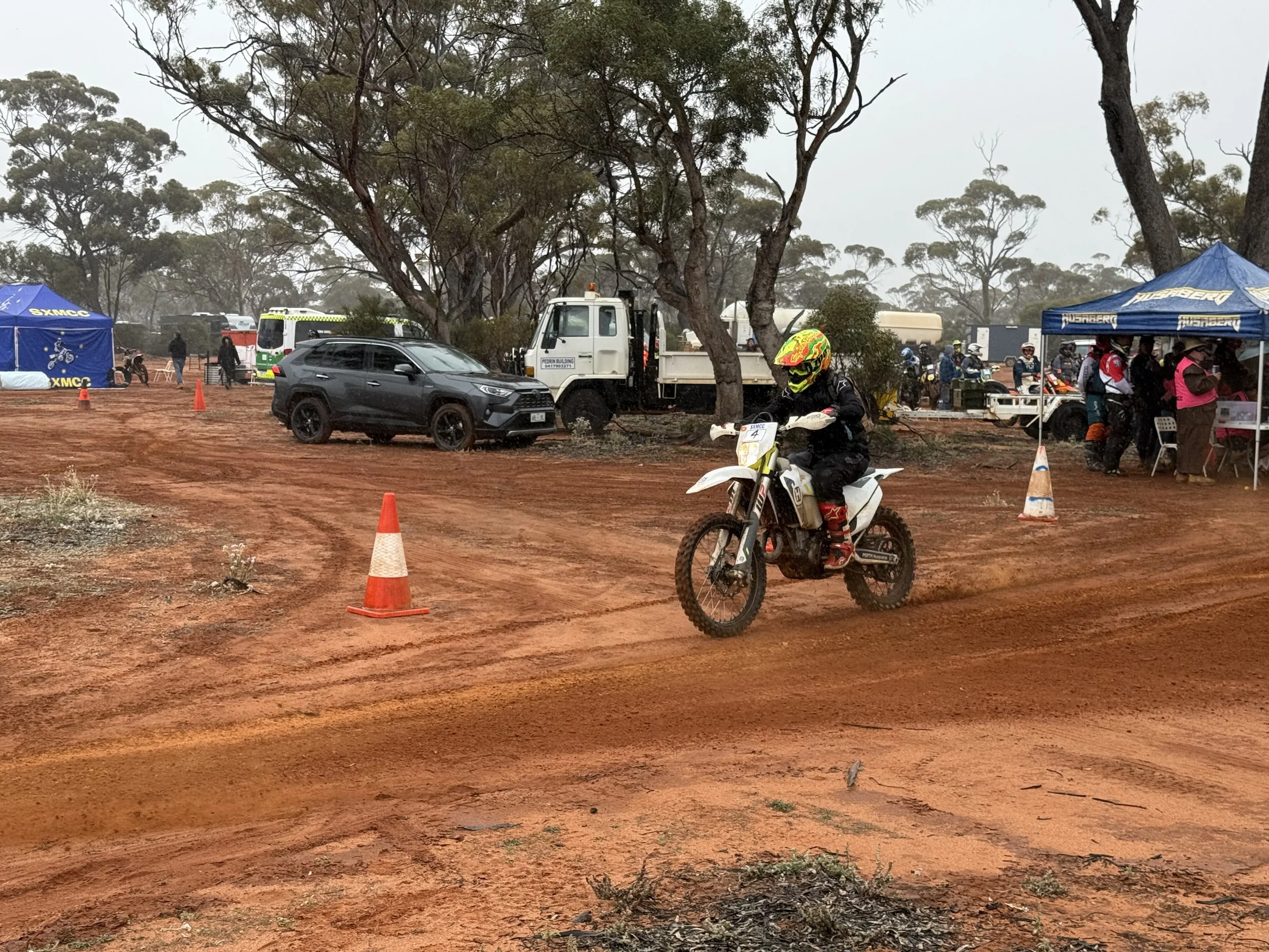 A person wearing a motorcycle helmet and riding gear on a dirt bike on a dirt track, with orange safety cones, trees, and tents in the background.