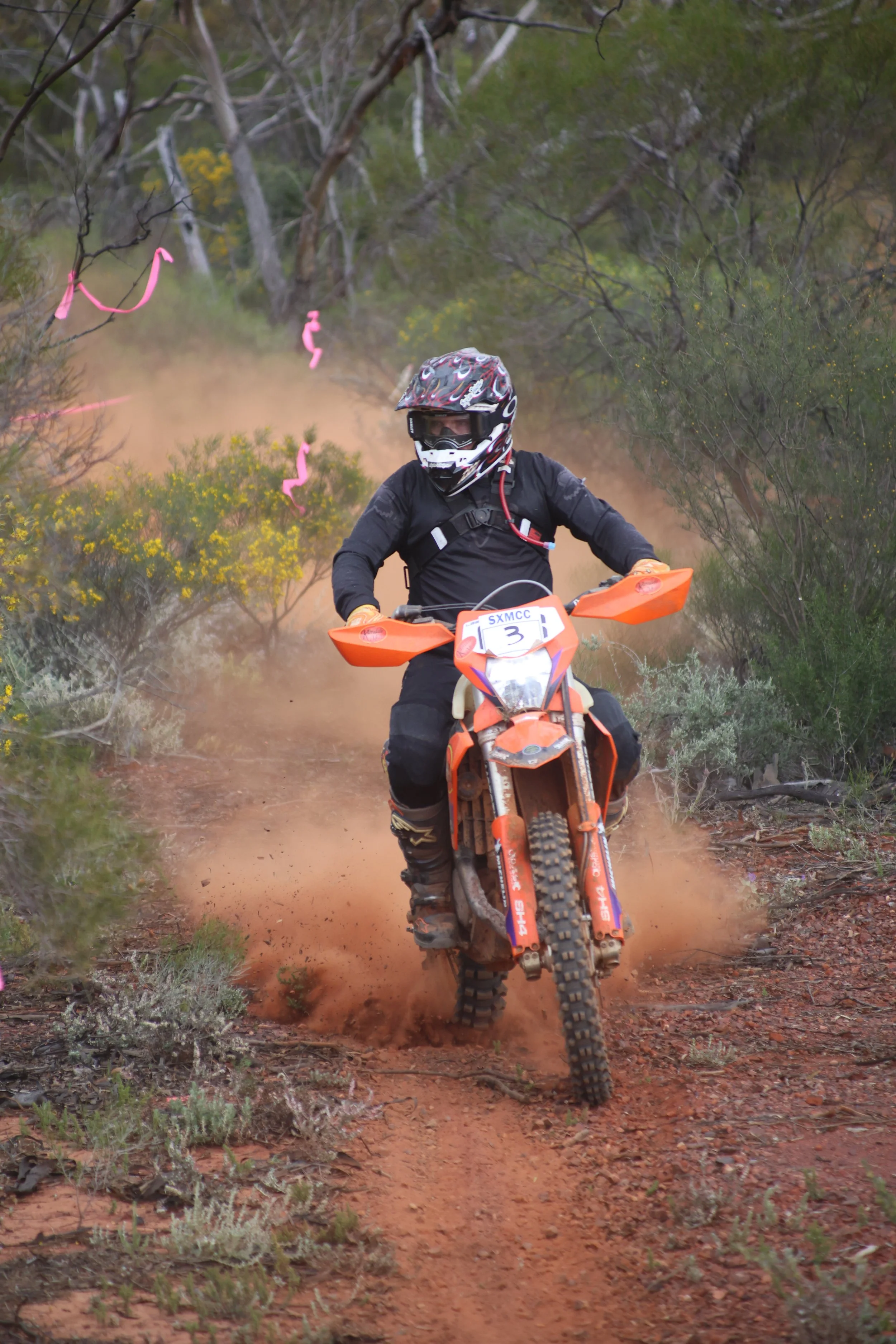 A person riding an off-road motorcycle through a dirt trail surrounded by bushes and trees, kicking up dust, wearing a black outfit, helmet, and gloves.