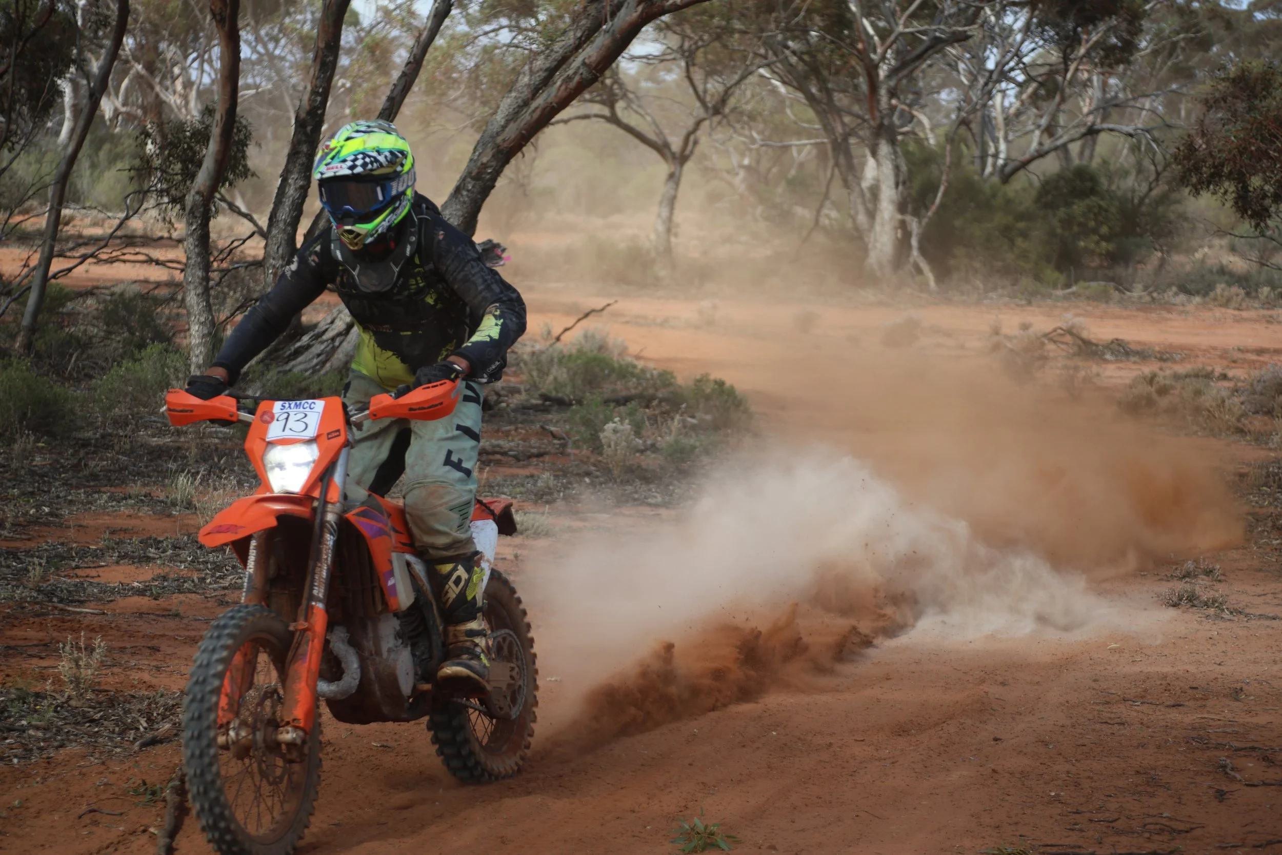 Motocross rider on an orange dirt bike racing through a dusty, wooded trail with trees and dry ground.