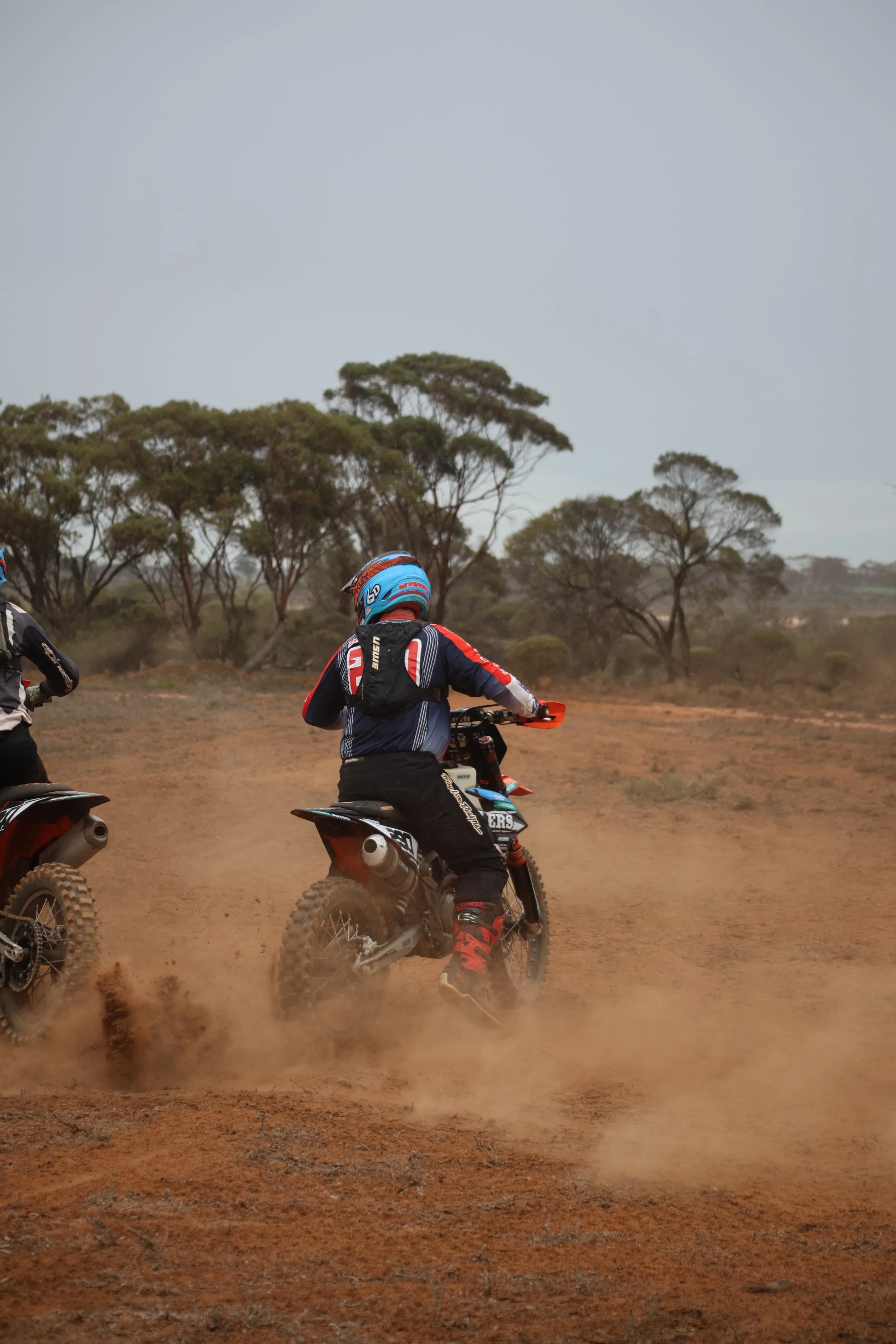 Two off-road motorcycle riders riding on dirt trail, kicking up dust, with sparse trees and a cloudy sky in the background.