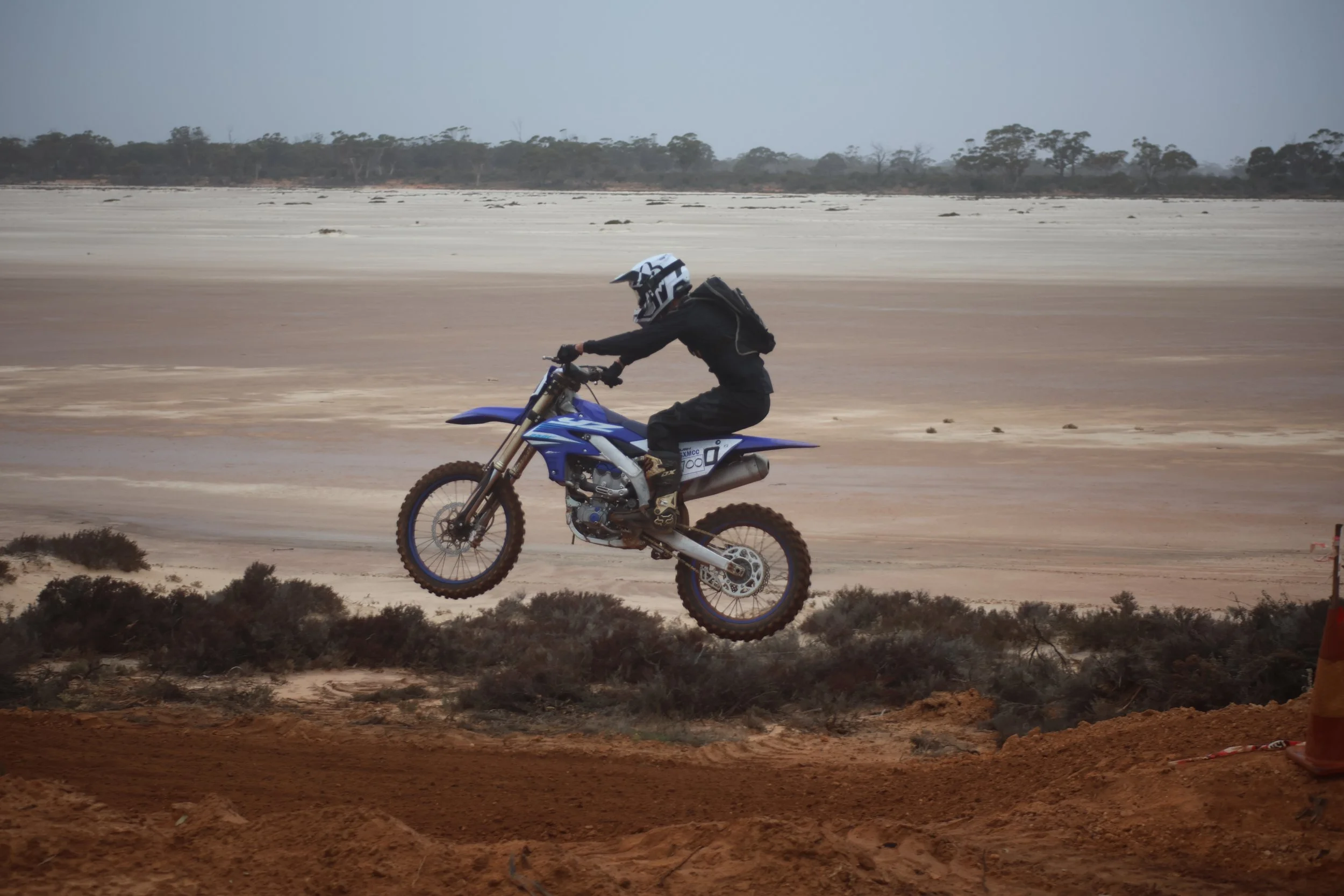 A person in black riding gear and a helmet jumping a dirt bike in a desert landscape with sparse vegetation and distant trees.