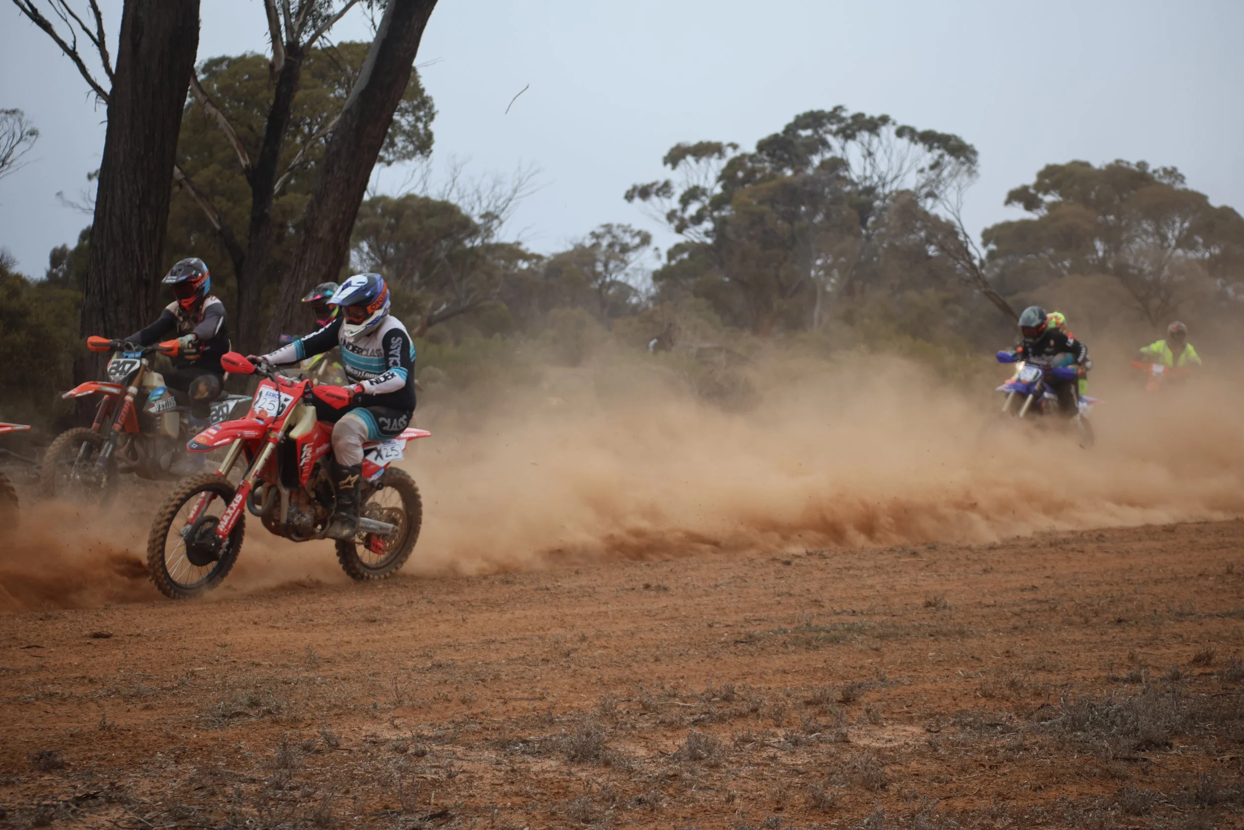 Motocross racers riding dirt bikes on a dusty trail in a wooded area.