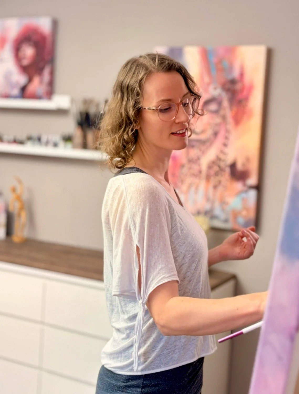 Eine Frau mit Brille, lockigen Haaren und im weißen T-Shirt, die vor einer Wand mit Gemälden in einem Kunststudio arbeitet.
