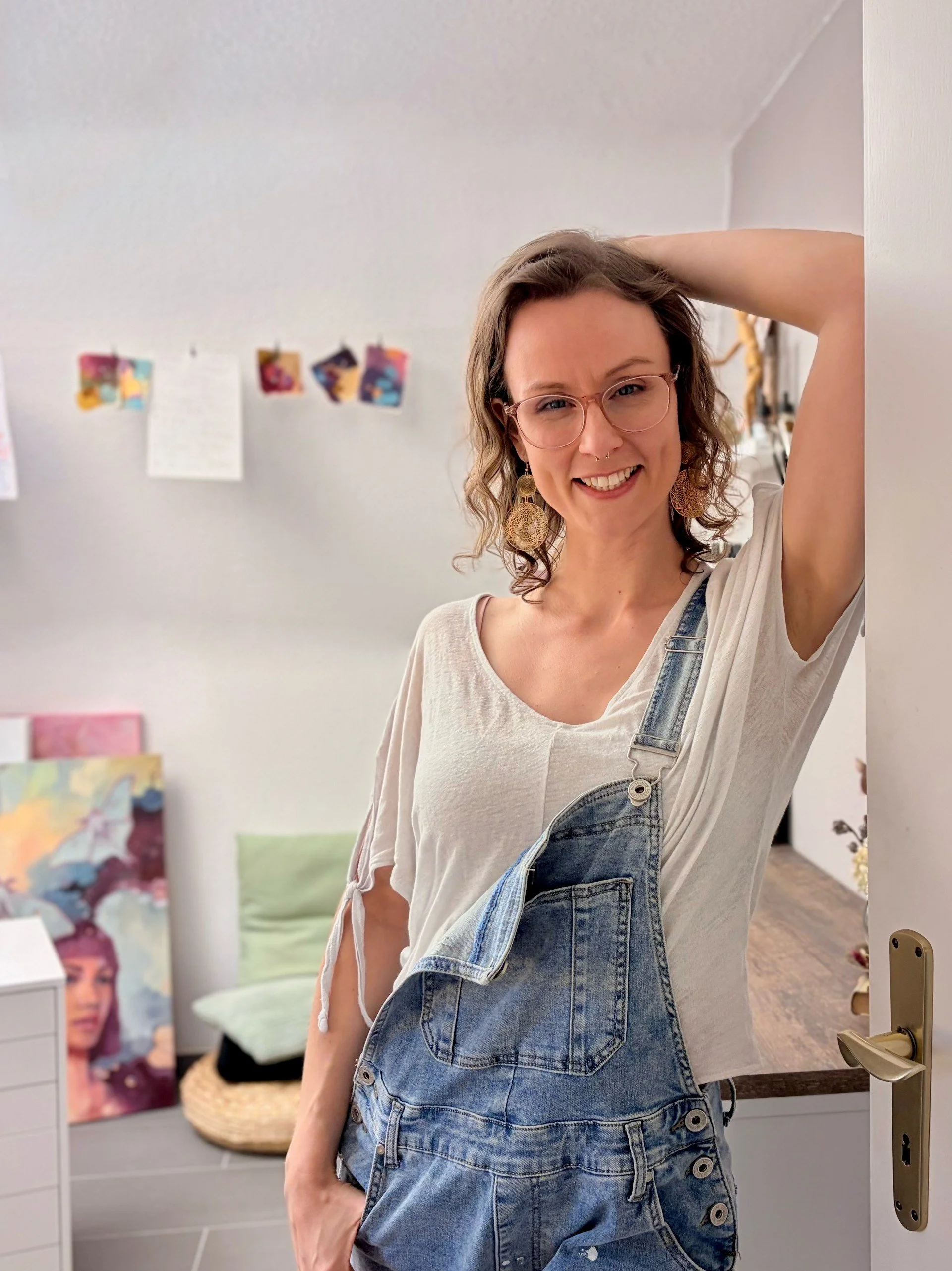 Junge Frau mit Brille, gelocktem Haar und großen Ohrringen, trägt ein weißes T-Shirt und Denim-Overall, steht in einem Raum mit Kunstwerken und Dekorationen im Hintergrund.