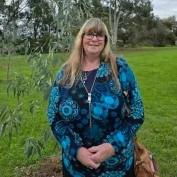 A woman with long blonde hair and glasses smiling outdoors in a grassy area with trees and tall plants, wearing a blue patterned jacket and carrying a brown purse.