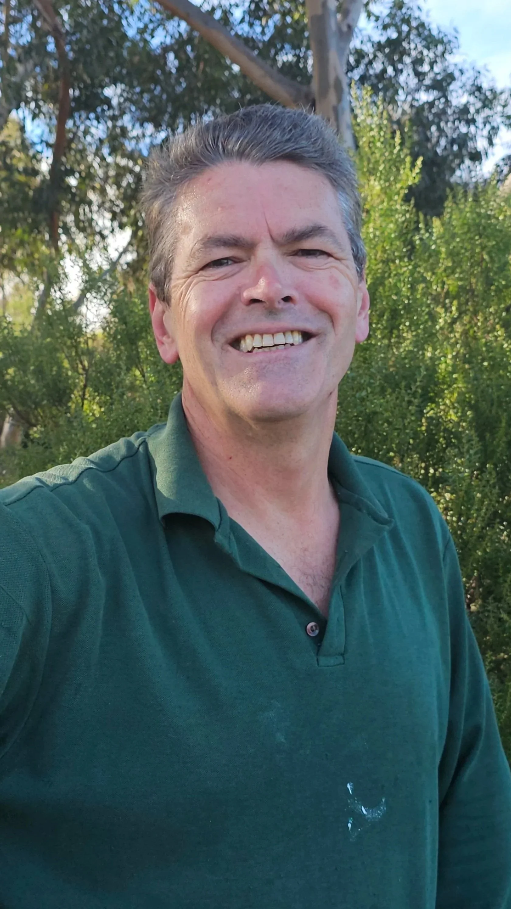 A smiling middle-aged man with gray hair in a green polo shirt taking a selfie outdoors with trees and blue sky in the background.