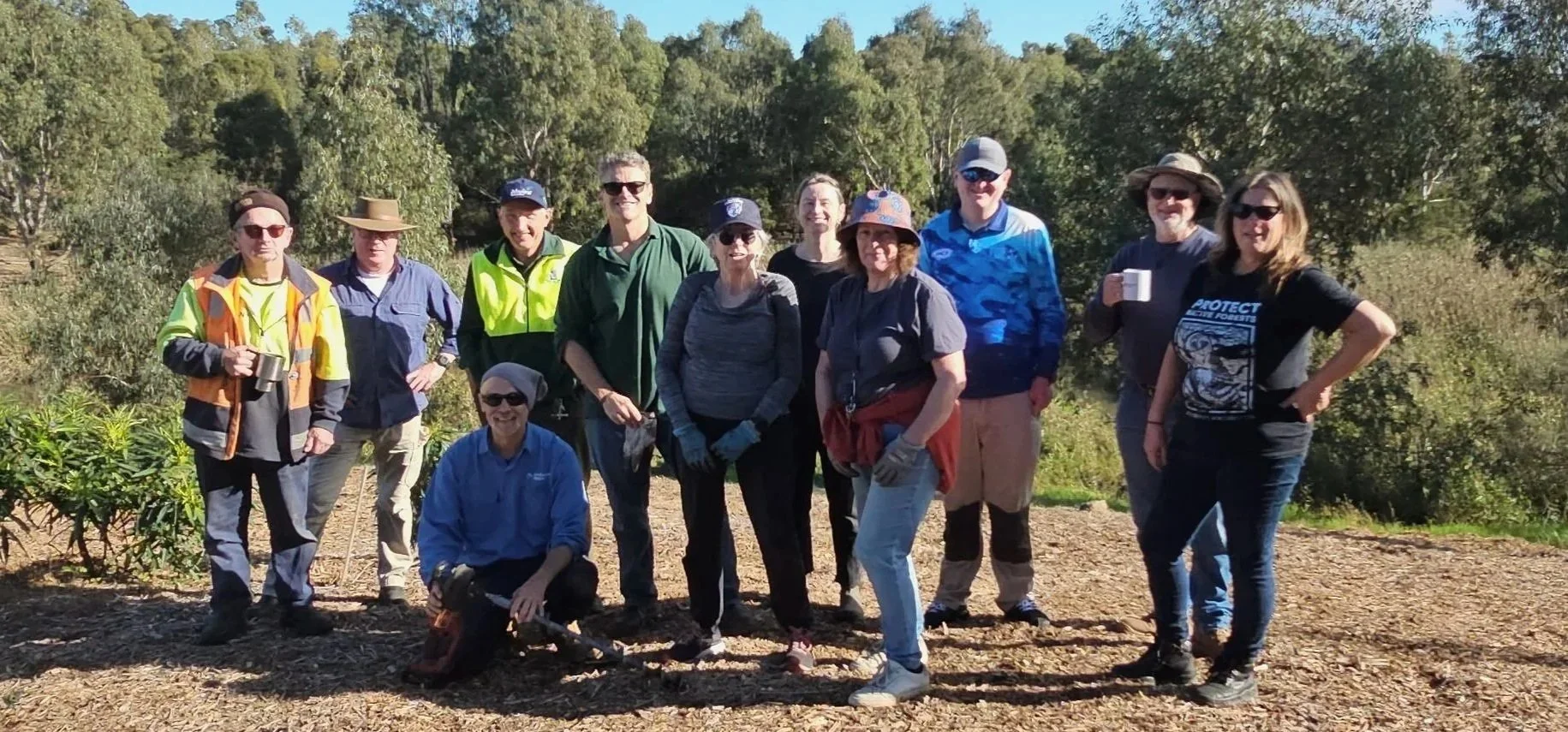 A group of eleven people standing outdoors on a trail surrounded by trees, wearing casual and outdoor clothing, some with hats and sunglasses, smiling for a photo on a sunny day.