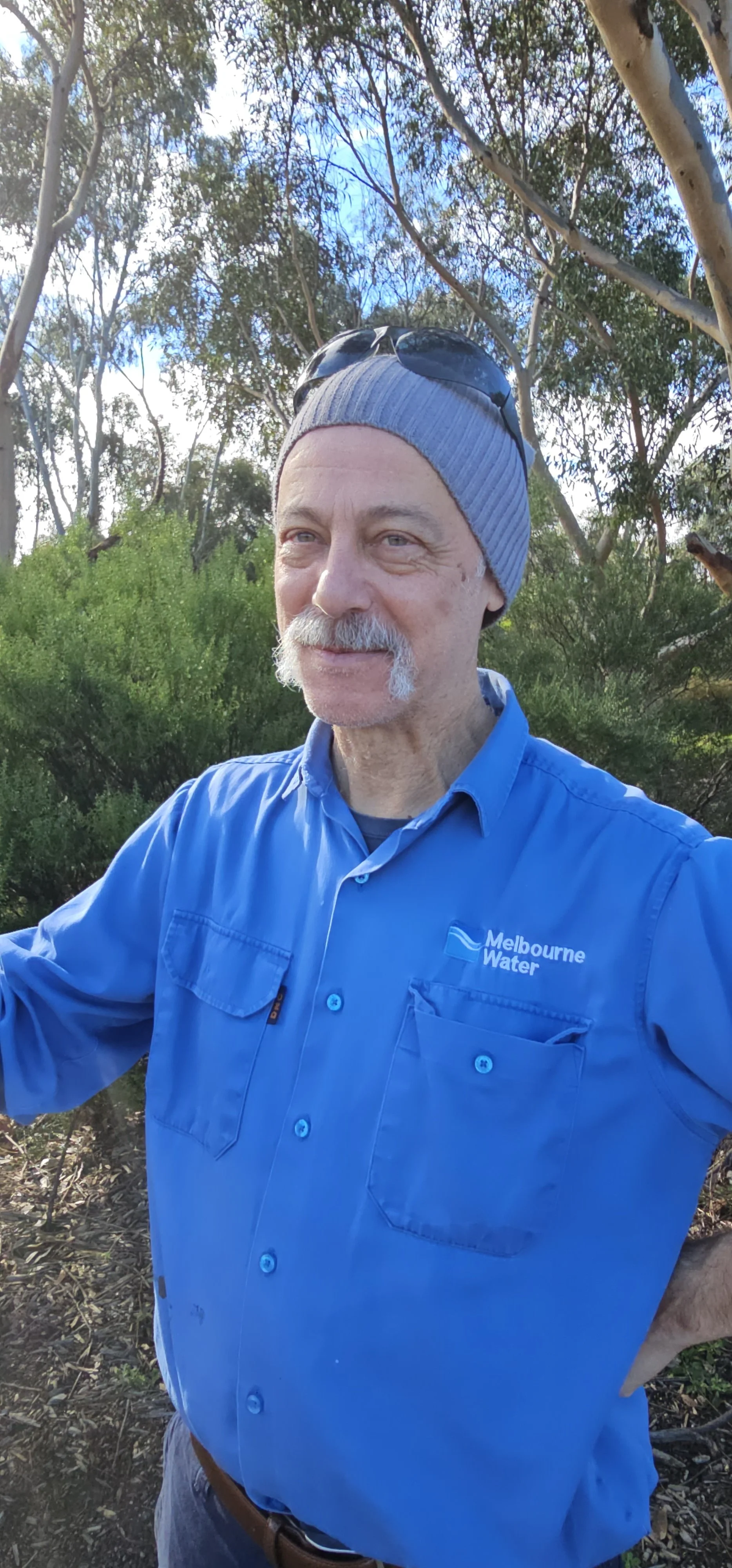 A man in a blue work shirt with the Melbourne Water logo stands outdoors with trees and blue sky in the background. He is wearing a gray beanie and sunglasses pushed up on his head. He has a mustache and light beard.