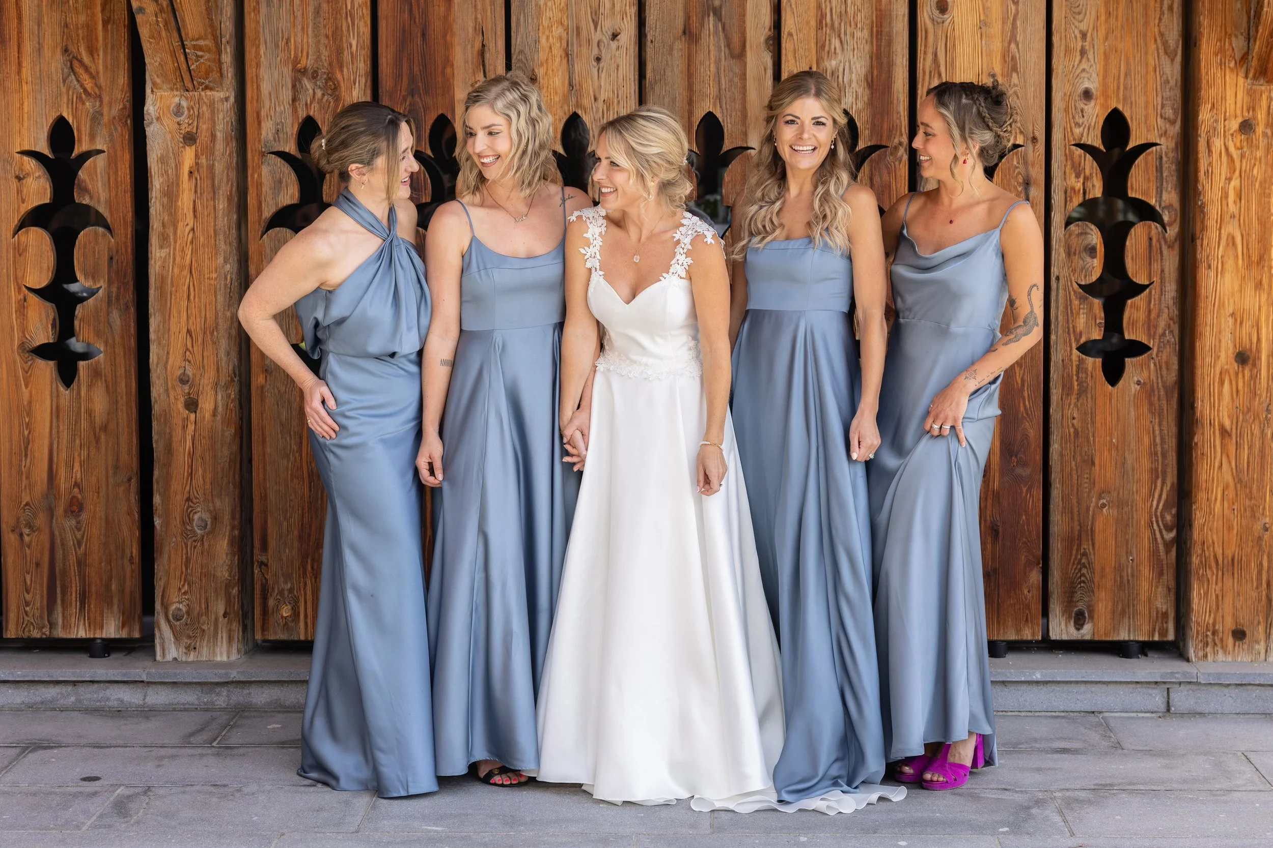 Bride and five bridesmaids standing together in front of a wooden fence, smiling and holding hands. The bride is wearing a white wedding gown with lace details on the shoulders, and the bridesmaids are in matching light blue dresses with various styl