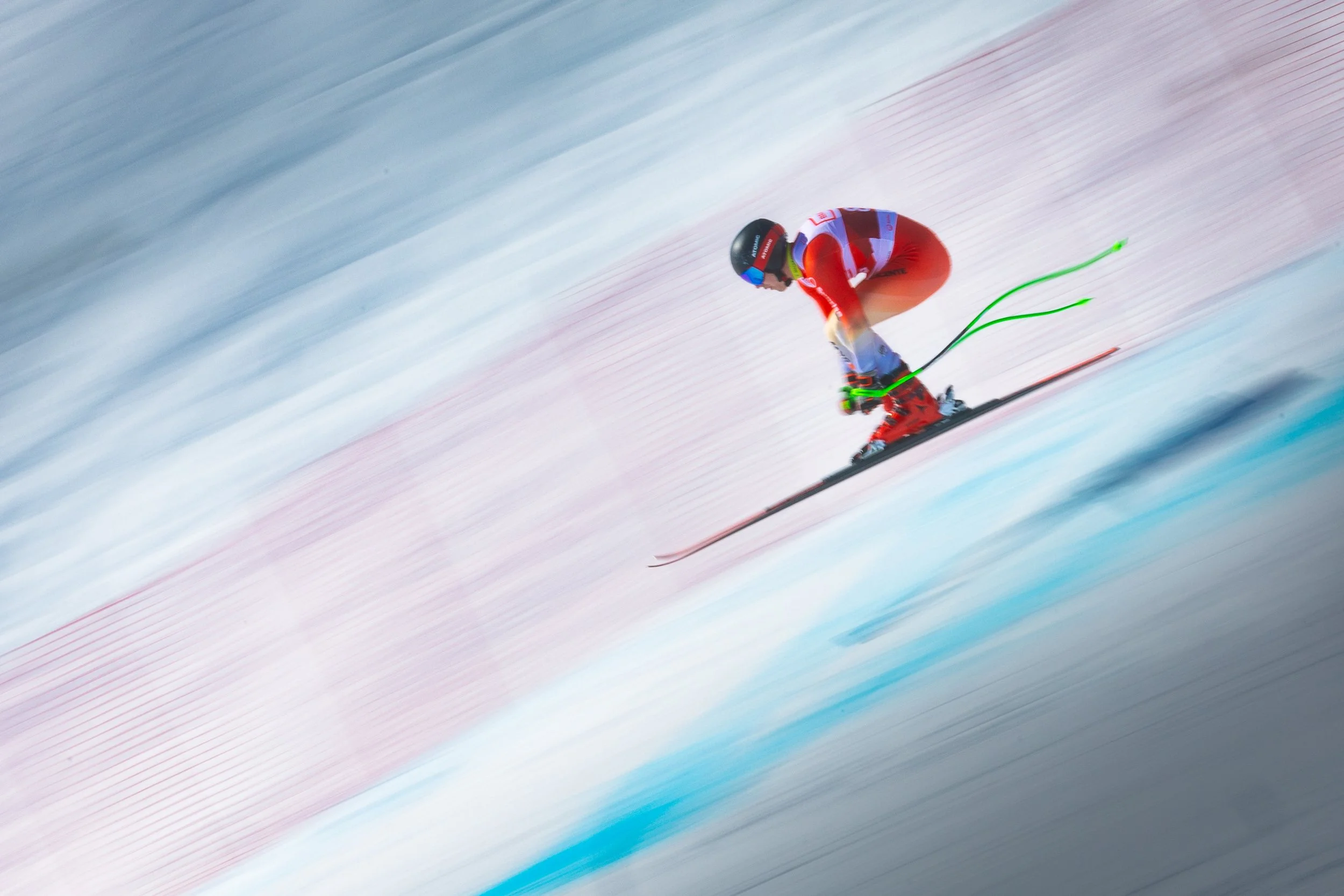 A skier dressed in red and black, wearing a helmet, is racing down a snowy slope with red protective fencing in the background.