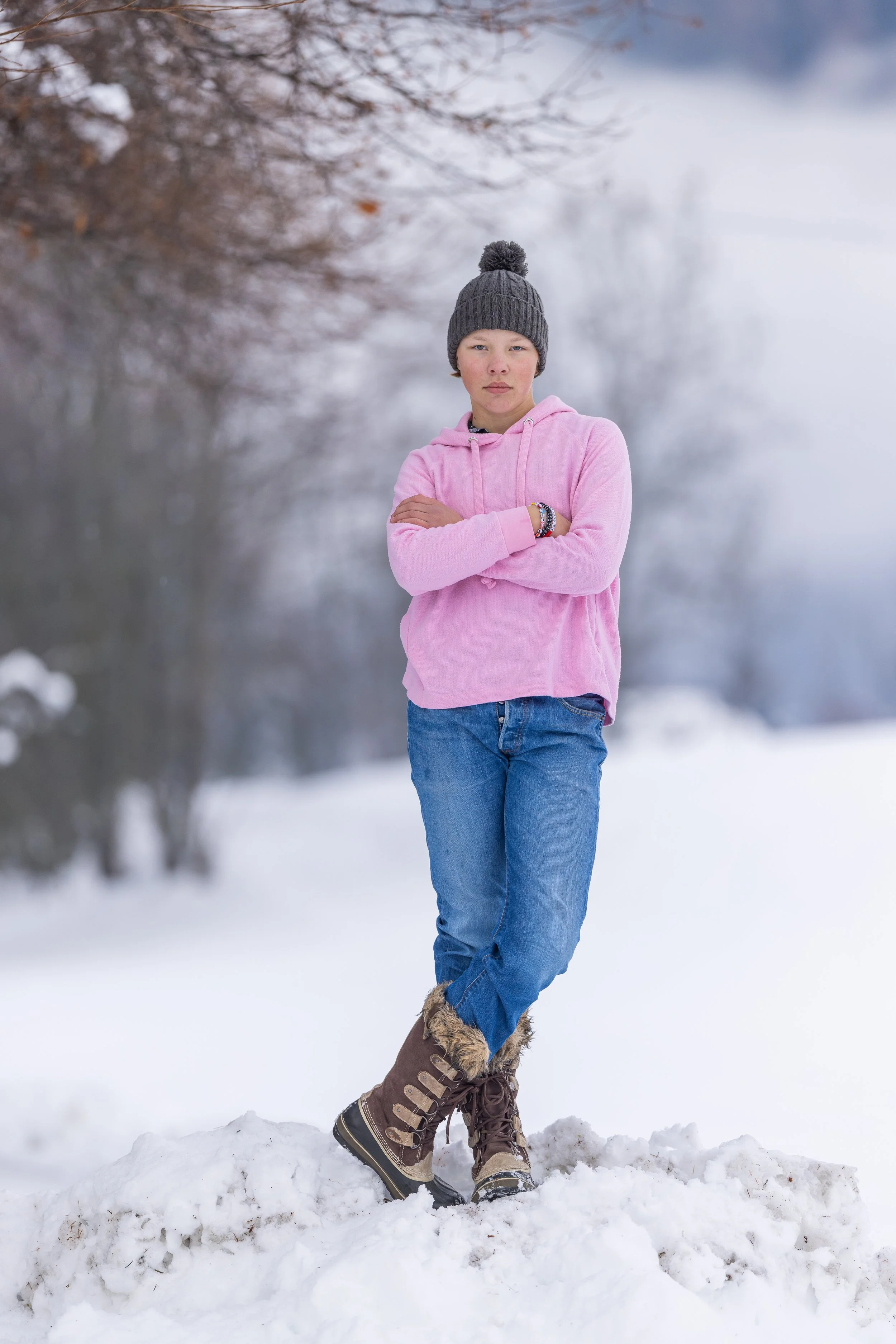 A girl standing on snow in a winter landscape, wearing a gray knit beanie, pink hoodie, blue jeans, and brown winter boots, with her arms crossed.