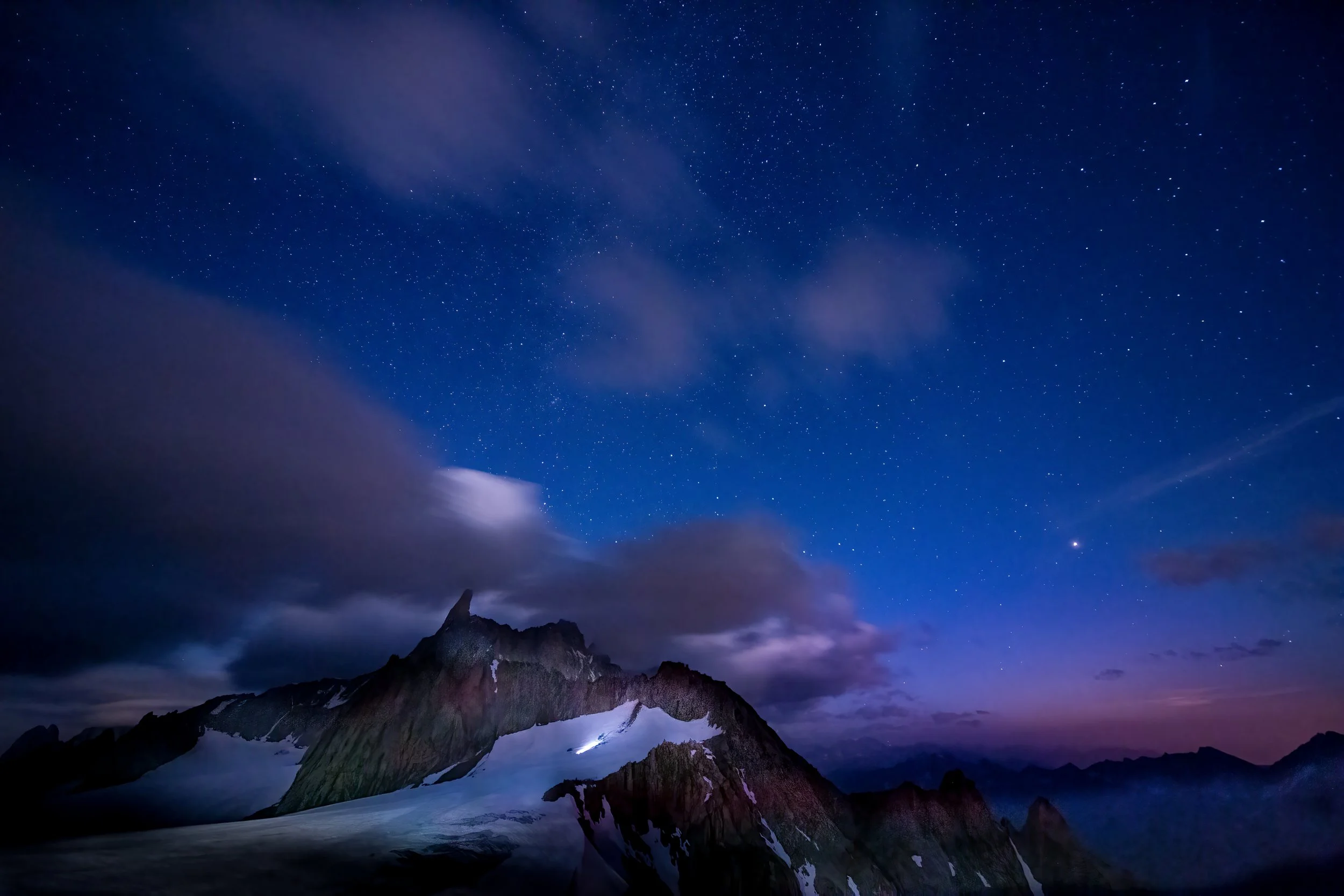 Nighttime landscape of a snow-capped mountain under a starry sky with clouds overhead.