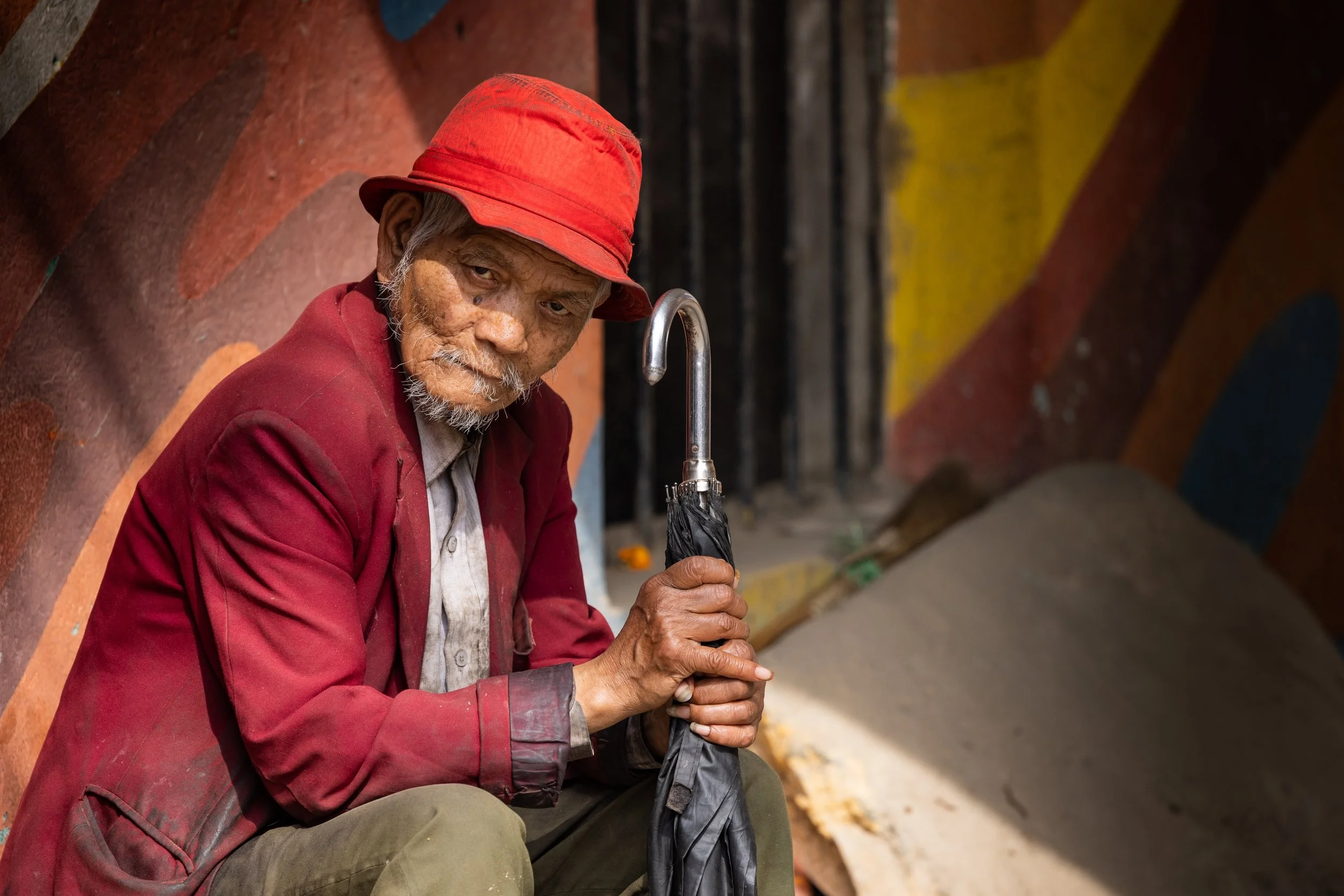 An elderly man with gray hair, beard, and weathered skin wearing a red hat and maroon jacket, holding a closed black umbrella, sitting against a colorful mural wall.