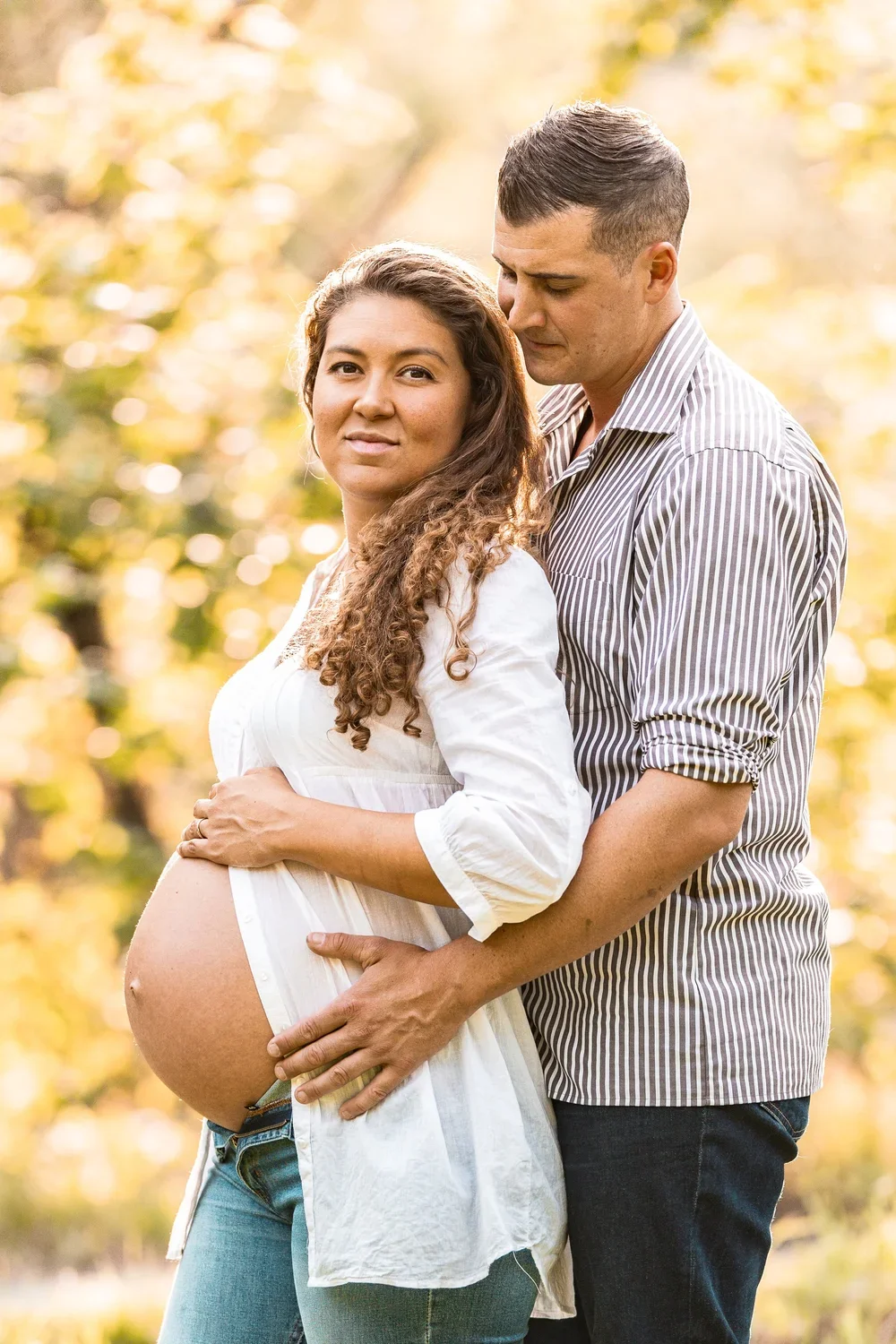 A pregnant woman with long curly hair and a white shirt stands outdoors with her partner. The man, wearing a striped shirt, stands behind her and gently holds her belly with both hands. They are in a sunlit, leafy area with a blurred background of yellow and green trees.