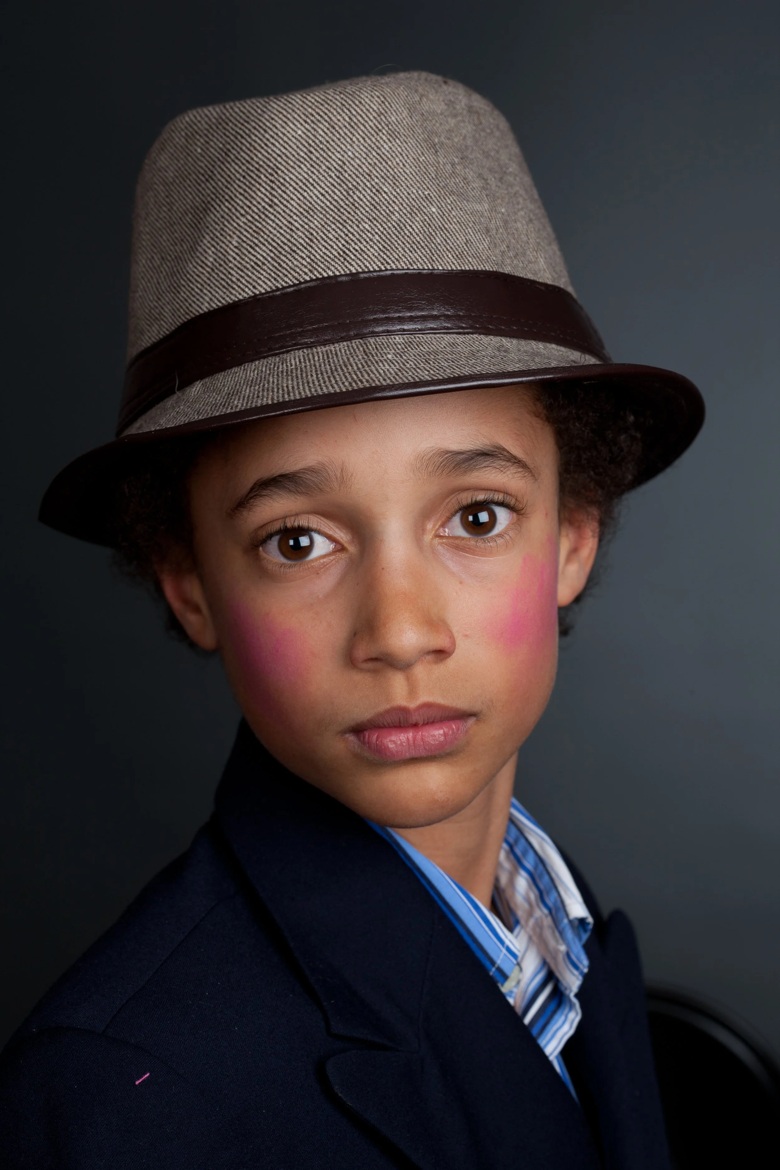 Portrait of a young person wearing a gray fedora hat with a brown band, a dark blazer, and a striped shirt, against a dark background.