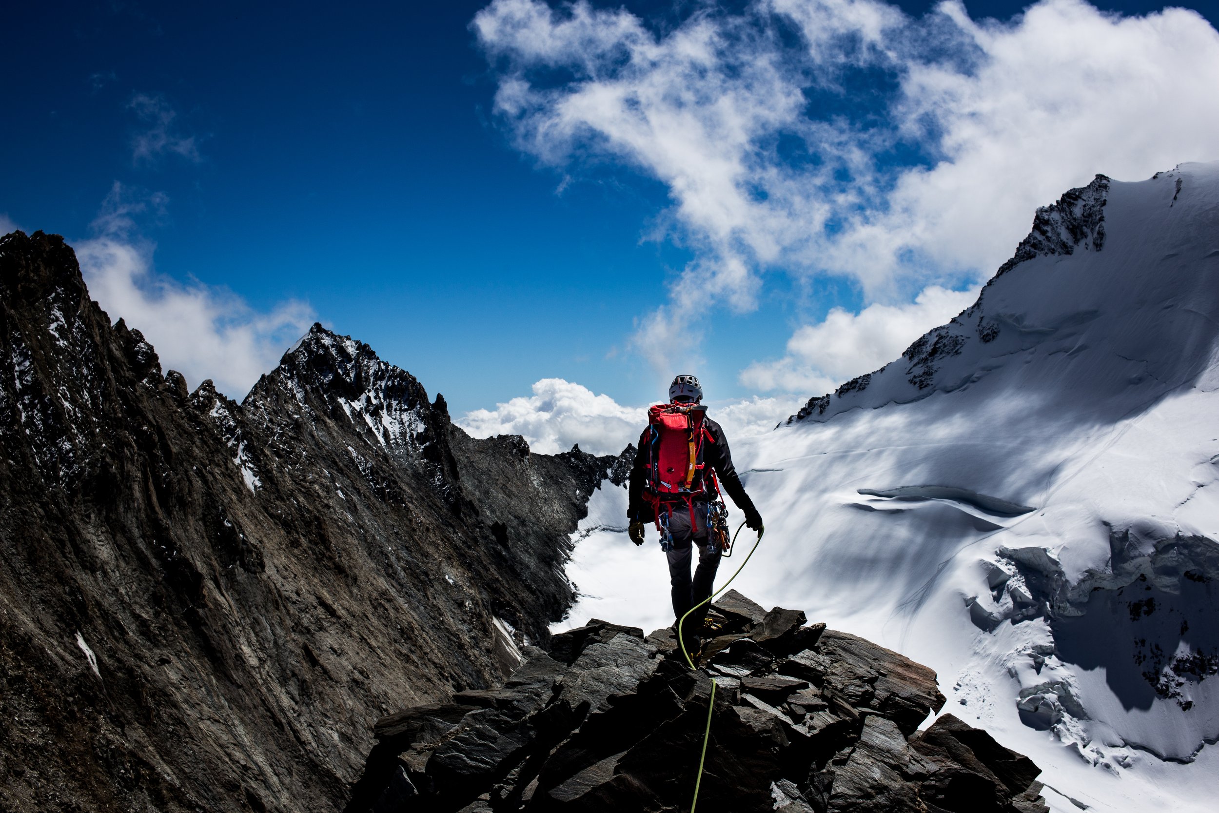 A mountain climber wearing a helmet, black clothing, and a red backpack walking on a rocky ledge in a snow-covered mountain range under a partly cloudy blue sky.