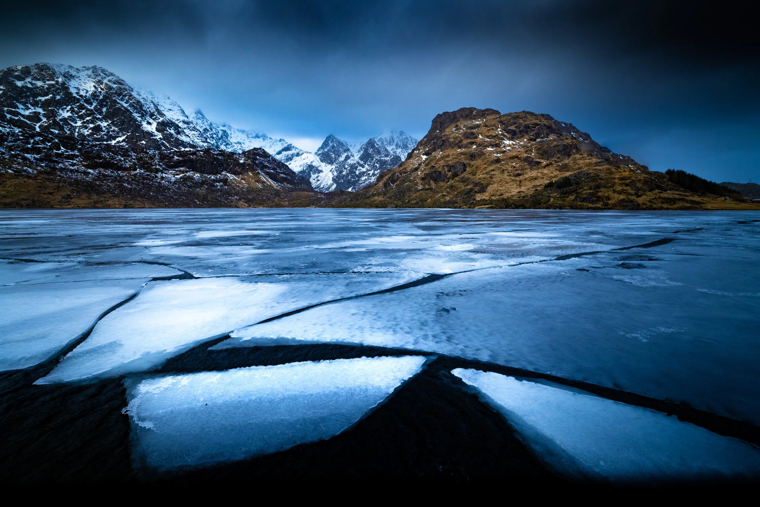Frozen lake with cracks in the ice, surrounded by snow-capped mountains under a dark cloudy sky.