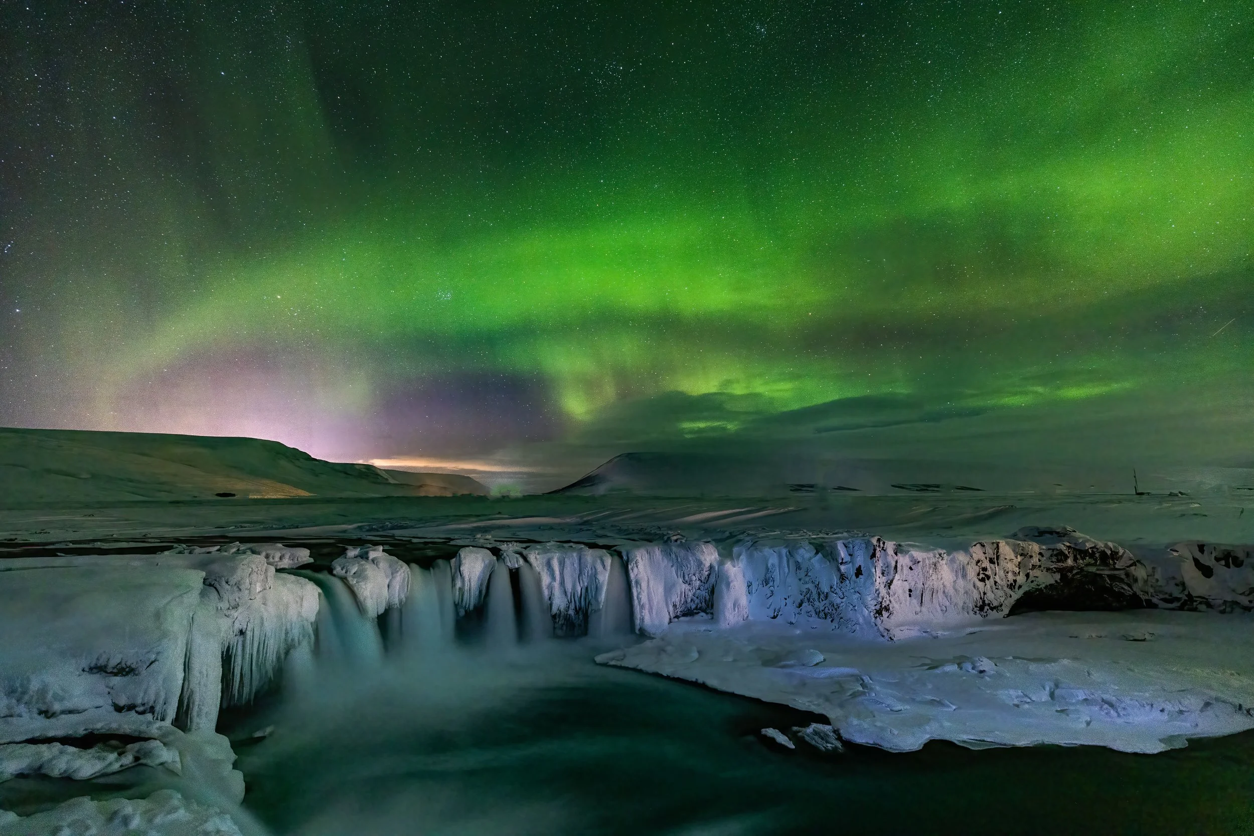 Northern lights illuminating a snowy landscape with a frozen waterfall and mountains in the background.