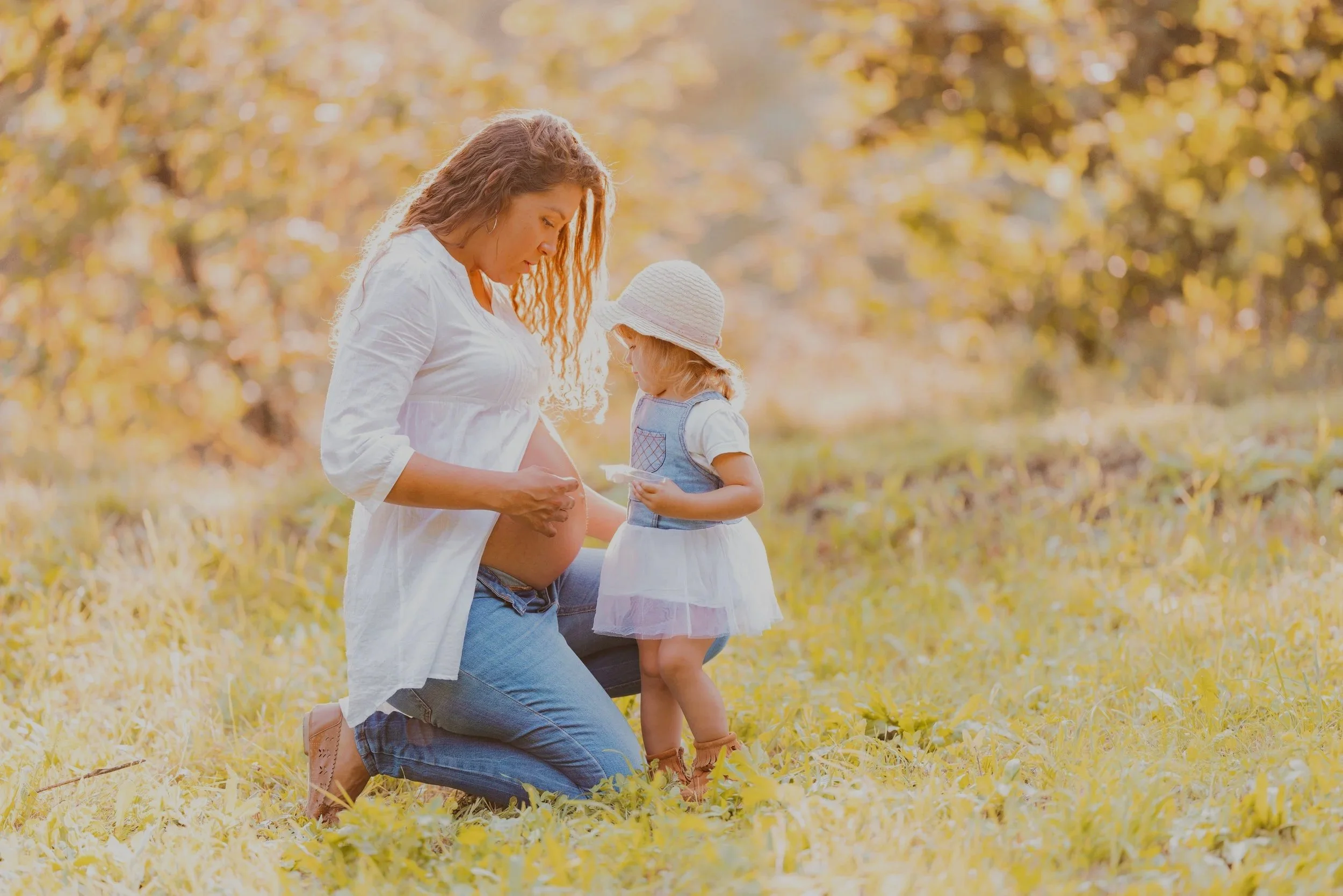 A pregnant woman in a white blouse and jeans kneeling in a field of yellow grass, looking at her young daughter who is wearing a white hat and light-colored dress, surrounded by trees and golden sunlight in Verbier Switzerland.